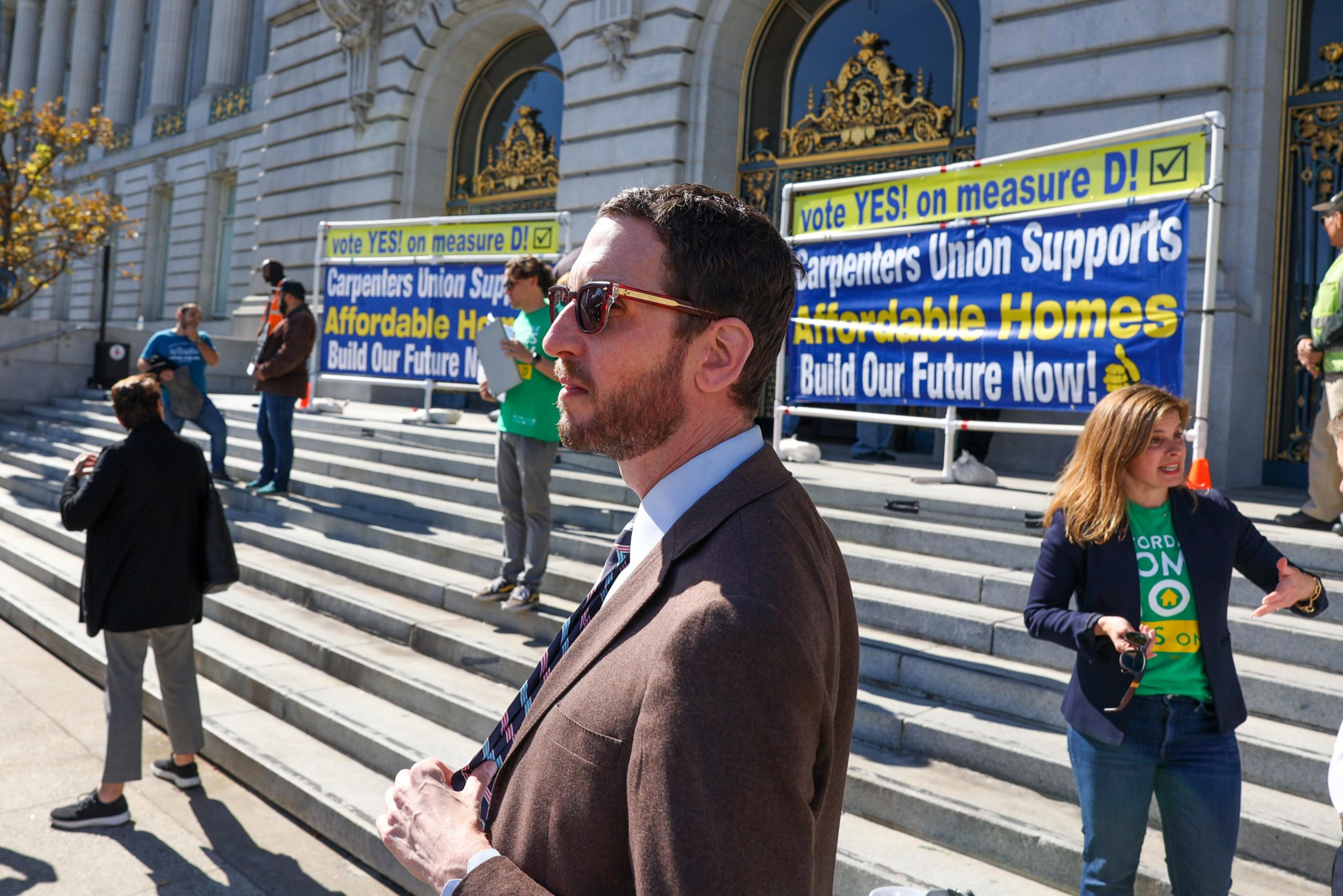 A rally on steps with a "Carpenters Union supports affordable homes" banner; people gathered, one in the foreground wearing a suit.