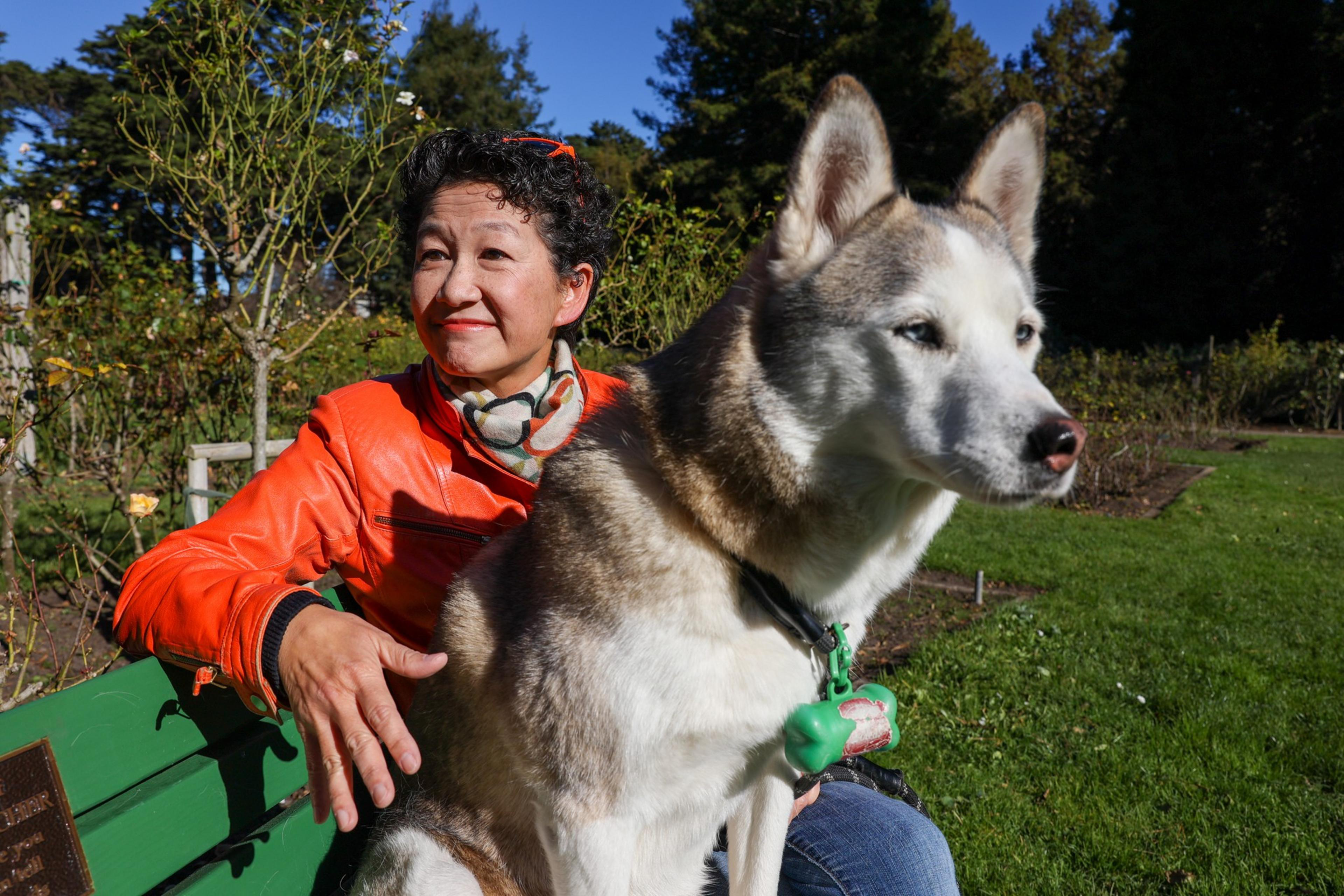 A woman in an orange jacket sits on a green bench outdoors with a large, light brown and white Siberian Husky in front of her.