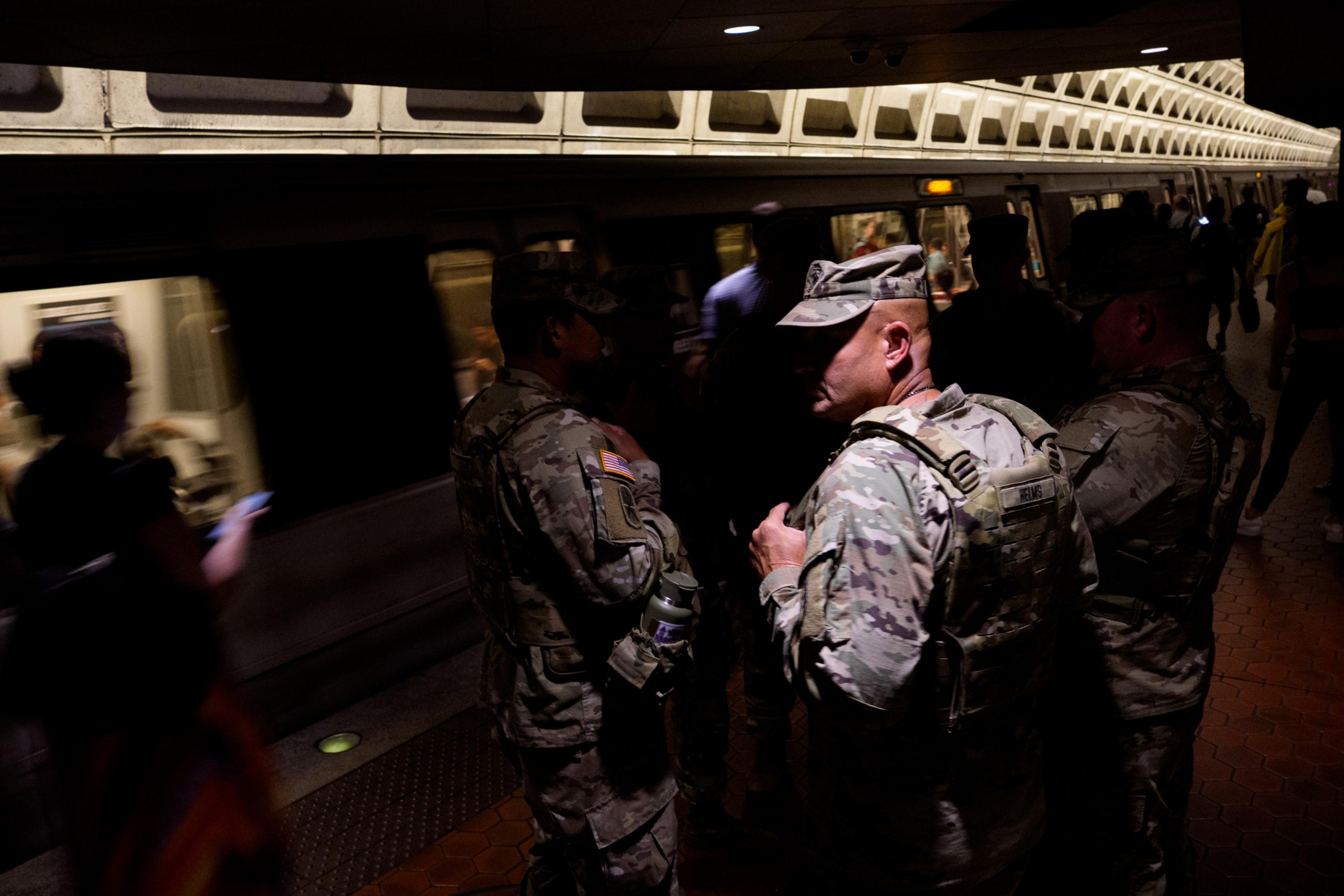 Several soldiers in camouflage uniforms stand on a dimly lit subway platform while a train passes by and other commuters wait nearby.