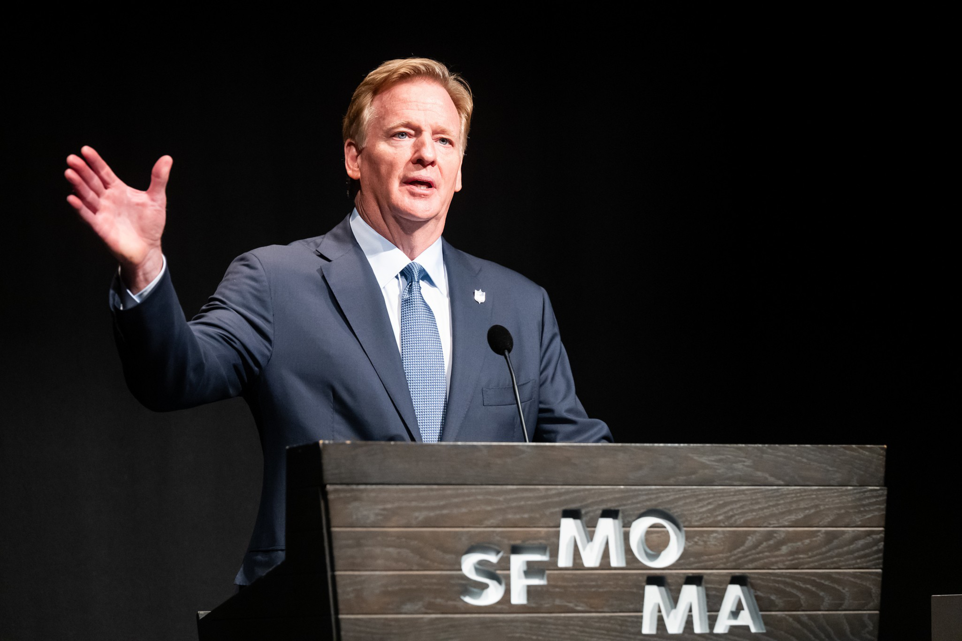 A man in a suit stands behind a podium labeled “SFMOMA,” speaking and gesturing with his right hand raised.