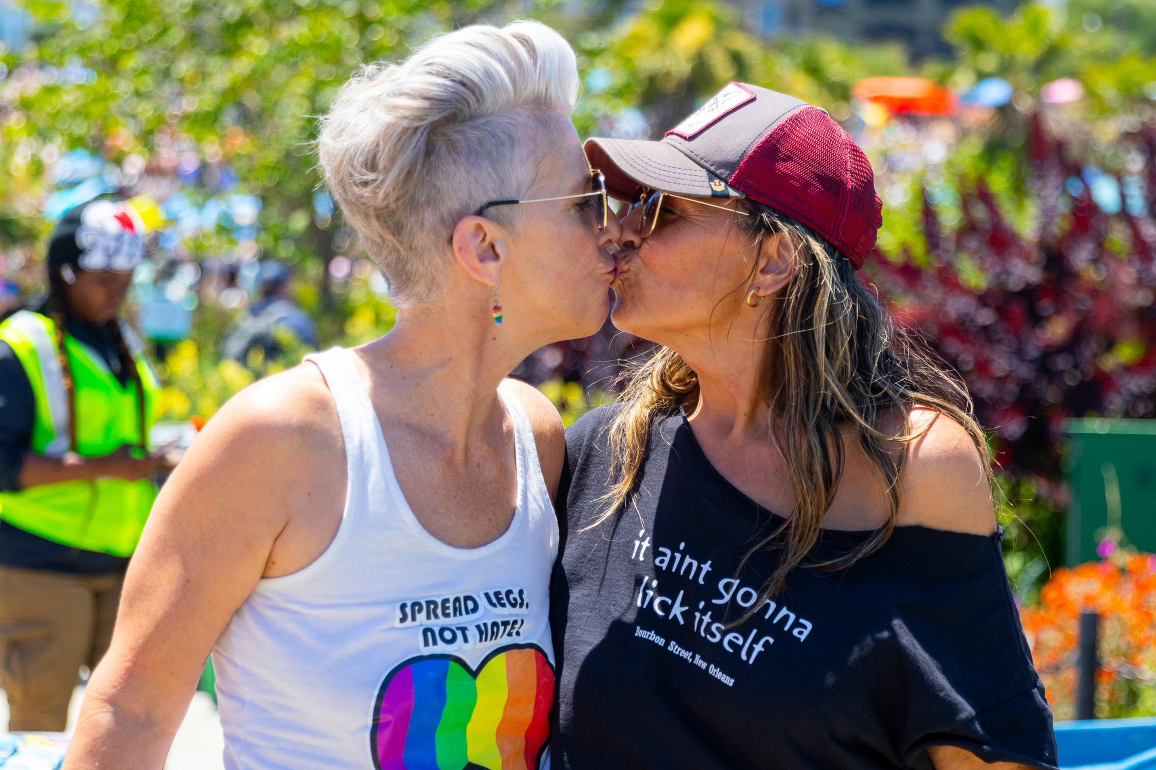 Two women wearing colorful outfits are kissing at an outdoor event. The woman on the left has short white hair, and the one on the right wears a hat and holds a rainbow fan.