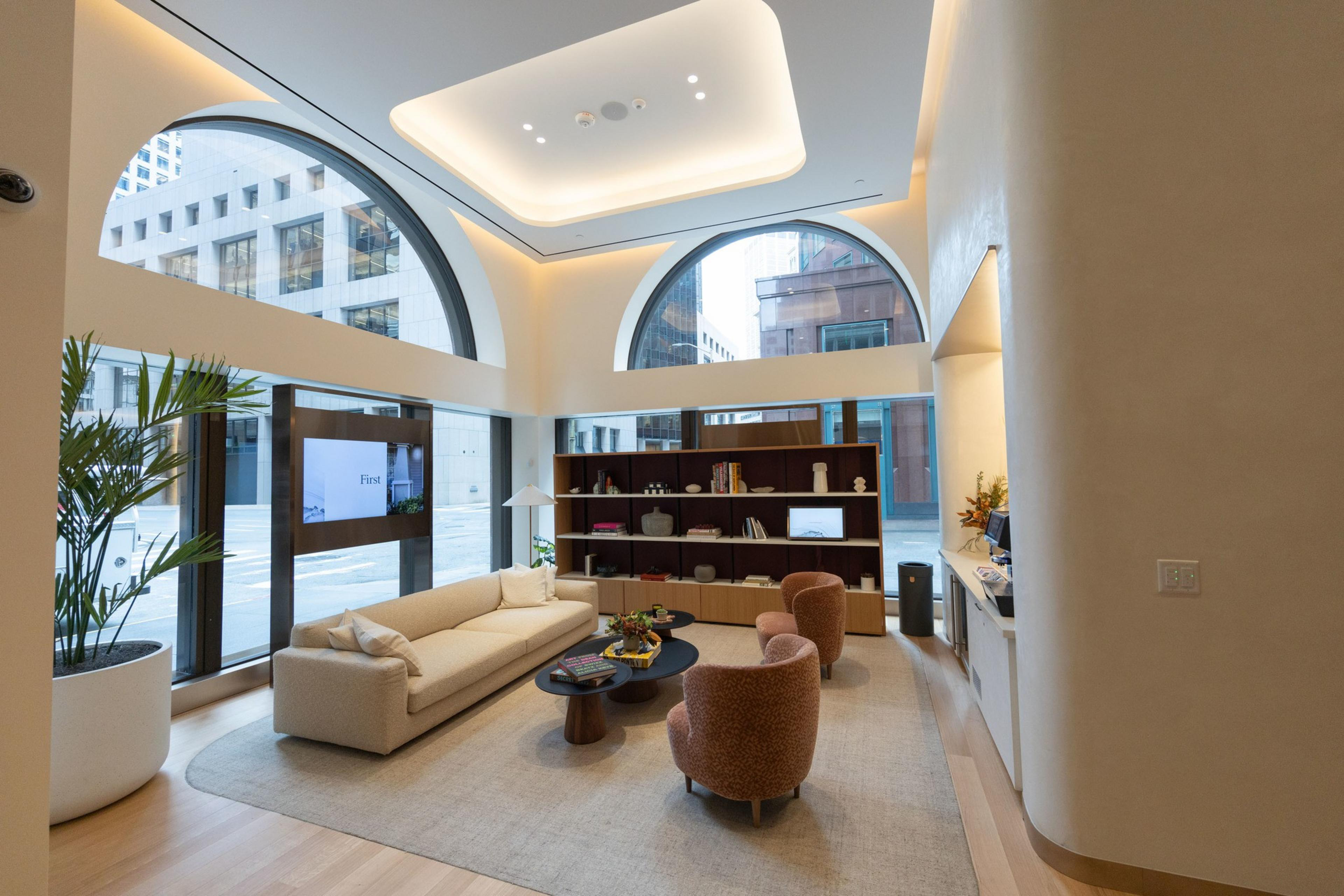 The image shows a modern, cozy lounge with large arched windows, soft lighting, plush seating, a beige couch, and two armchairs around a coffee table.