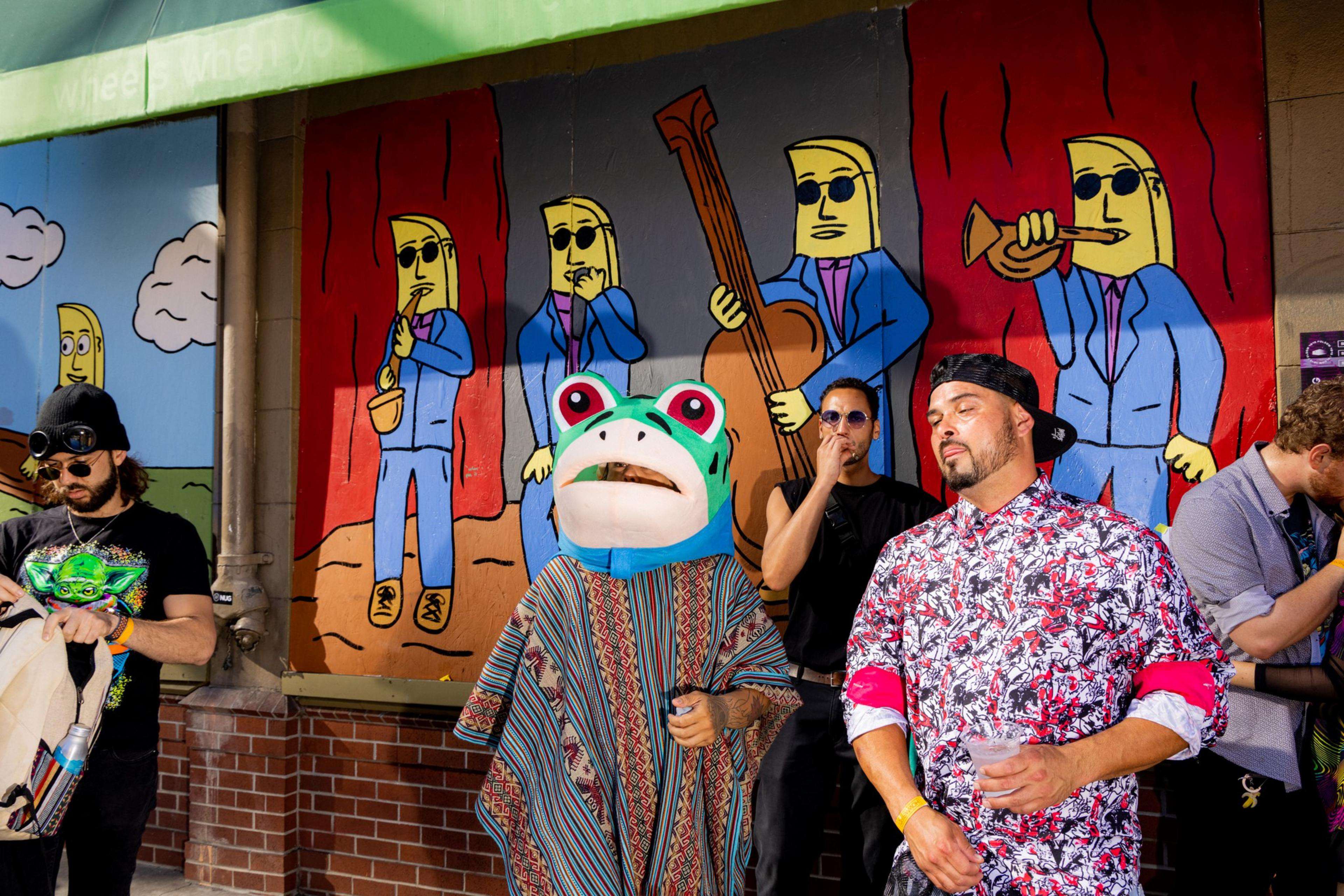 People in colorful outfits, including a frog mask, pose in front of a whimsical mural of yellow characters in blue suits with musical instruments.