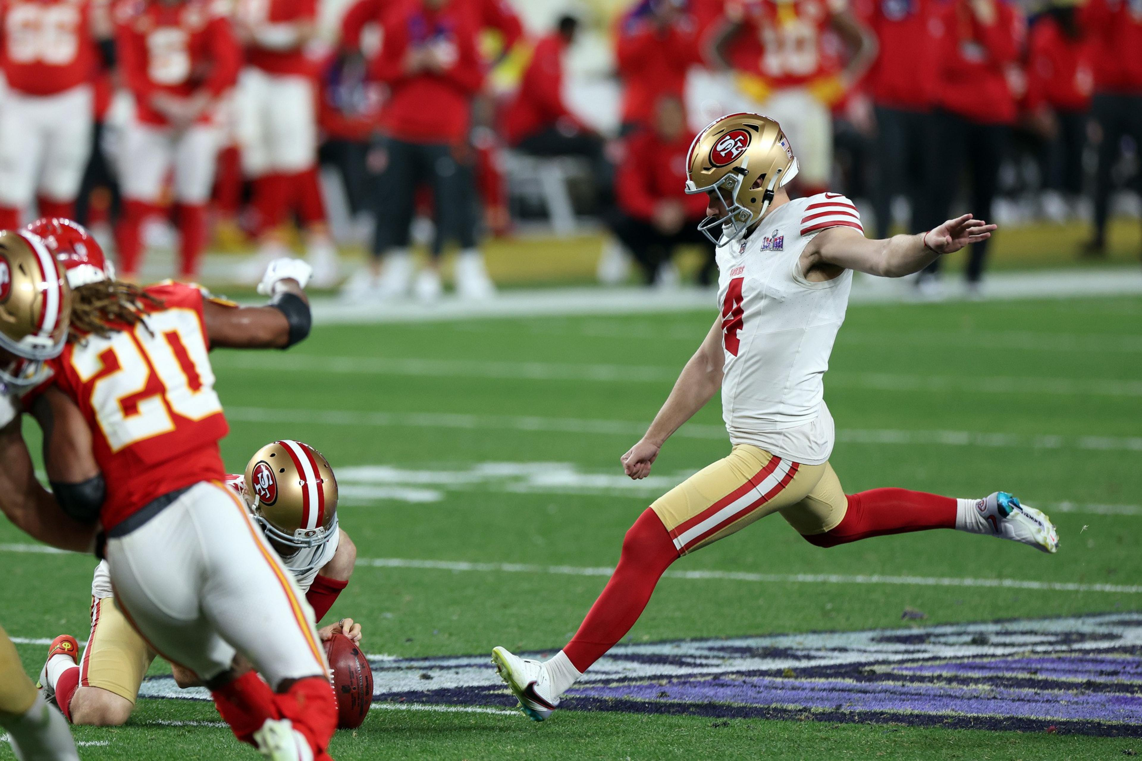 A football kicker is mid-kick on field with players around, in a professional game.