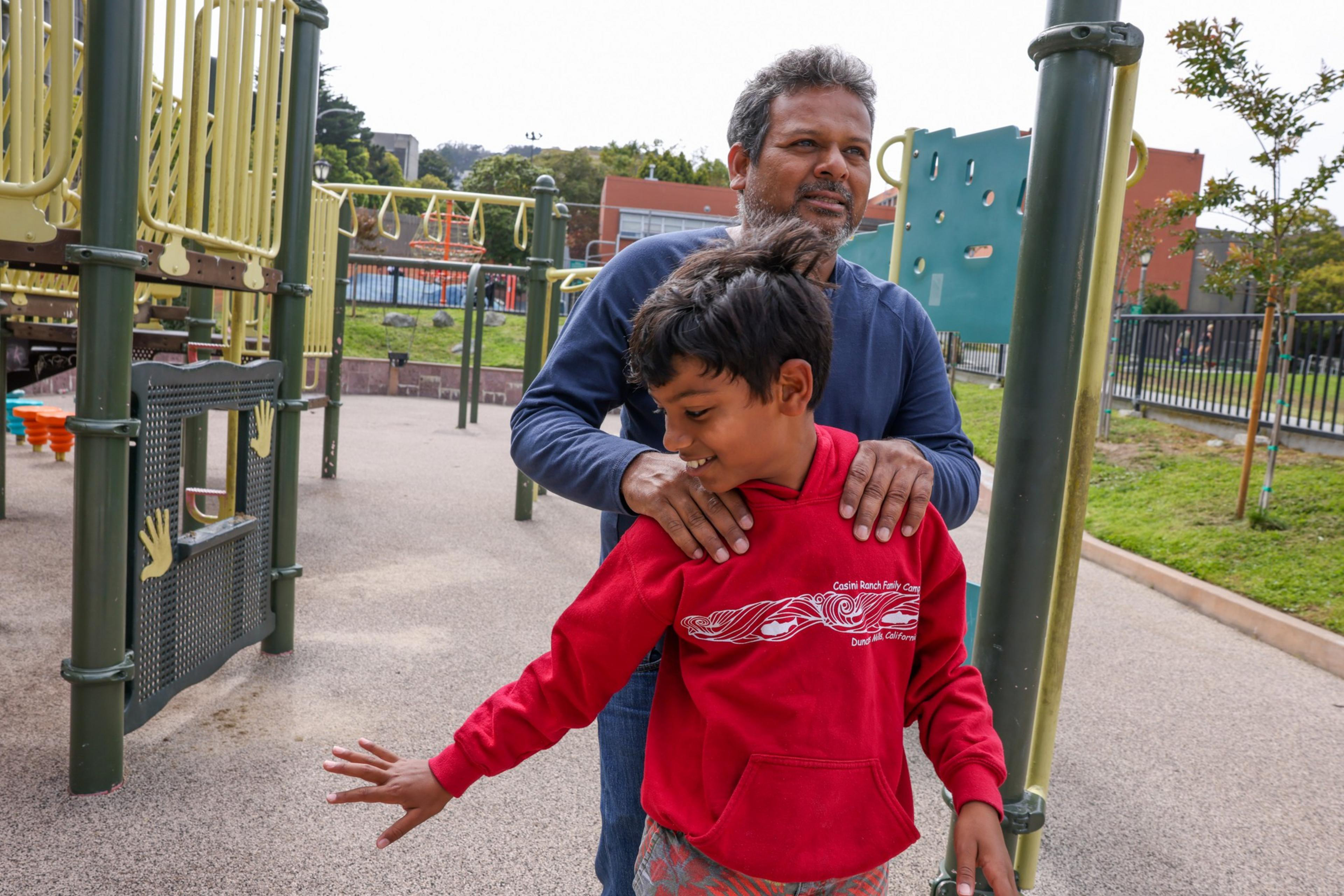A man stands behind a smiling boy in a red hoodie at a playground, gently holding his shoulders as the boy stretches his arm out.