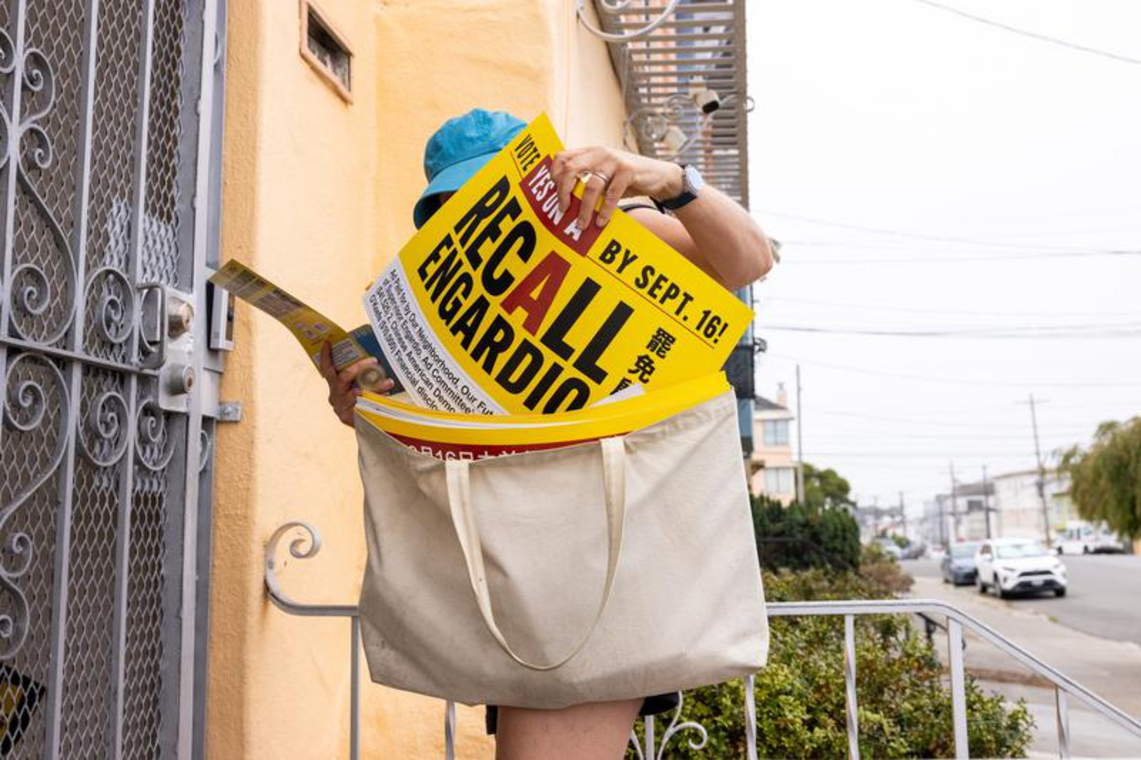 A person wearing a blue cap holds yellow "Recall Engardio" signs while packing them into a large beige tote bag by a yellow building.