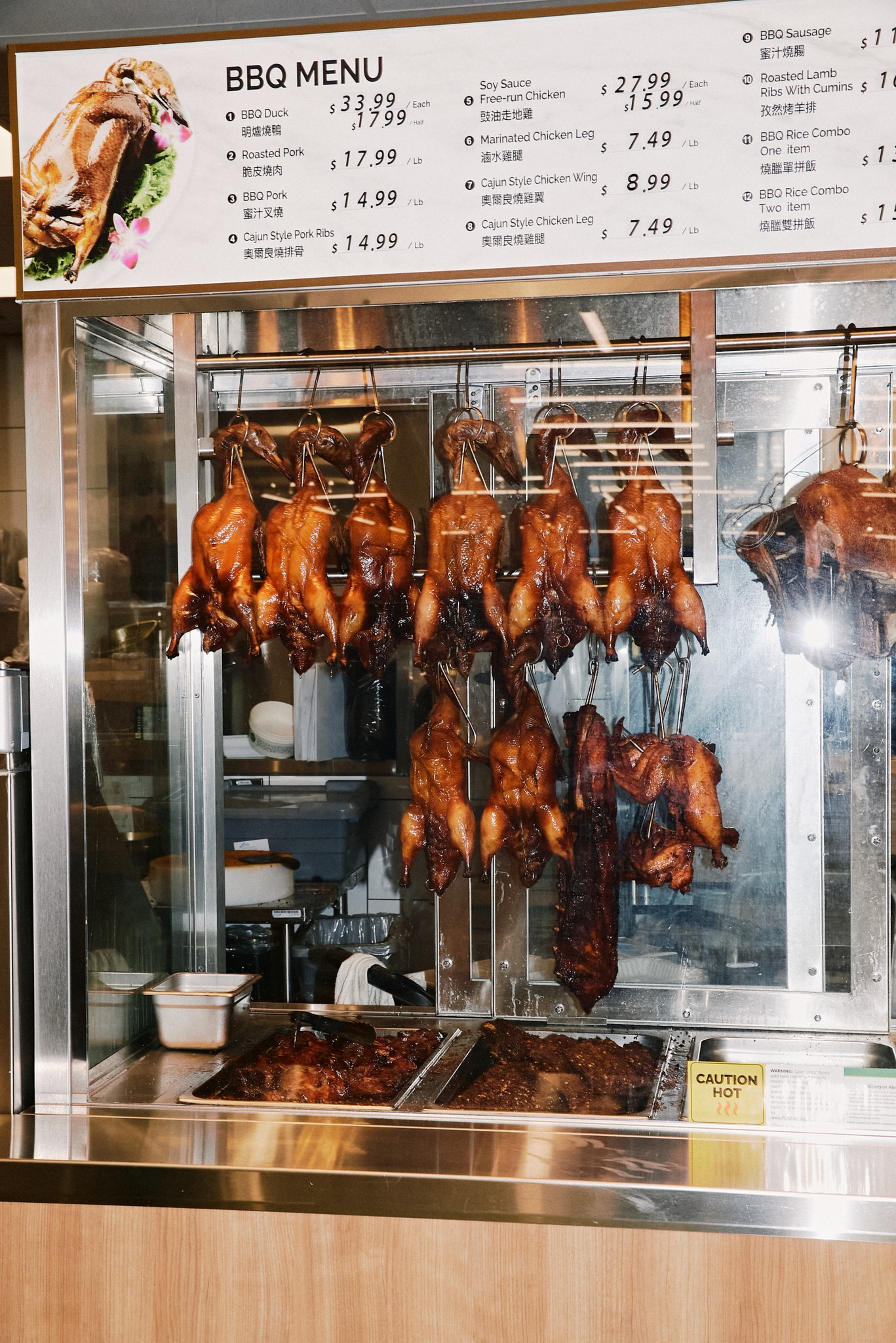 Roasted whole ducks and chicken hanging on hooks inside a glass display, with trays of cooked meat below and a BBQ menu above.