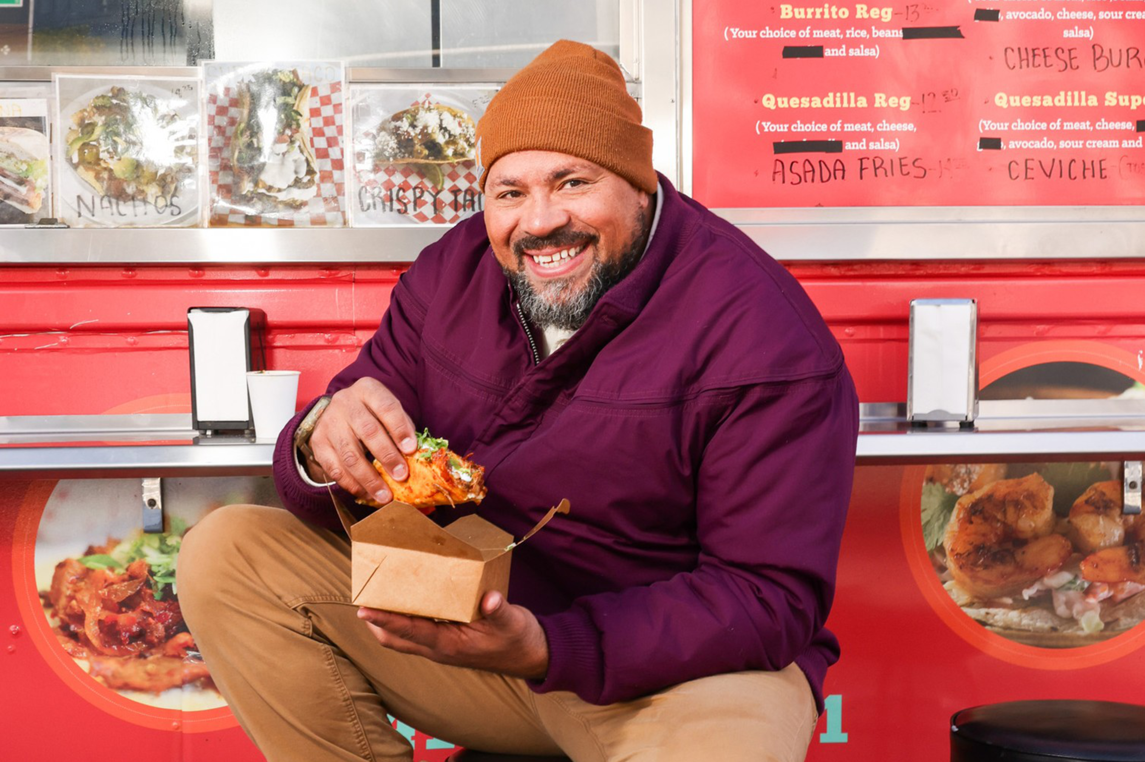 A man in a purple jacket and brown beanie joyfully holds a taco while sitting on a stool in front of a red food truck with menu items displayed.