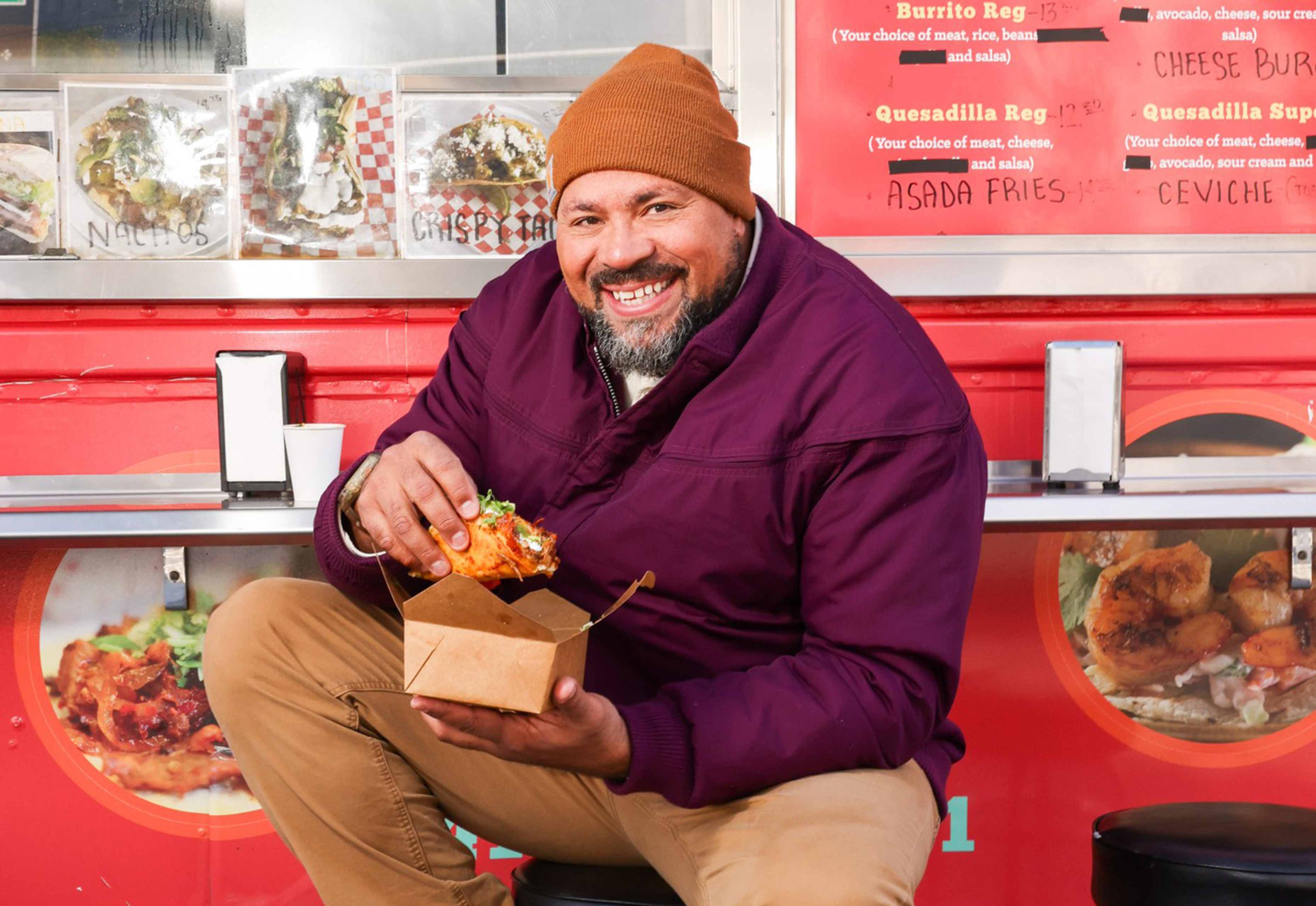 A man in a purple jacket and brown beanie joyfully holds a taco while sitting on a stool in front of a red food truck with menu items displayed.