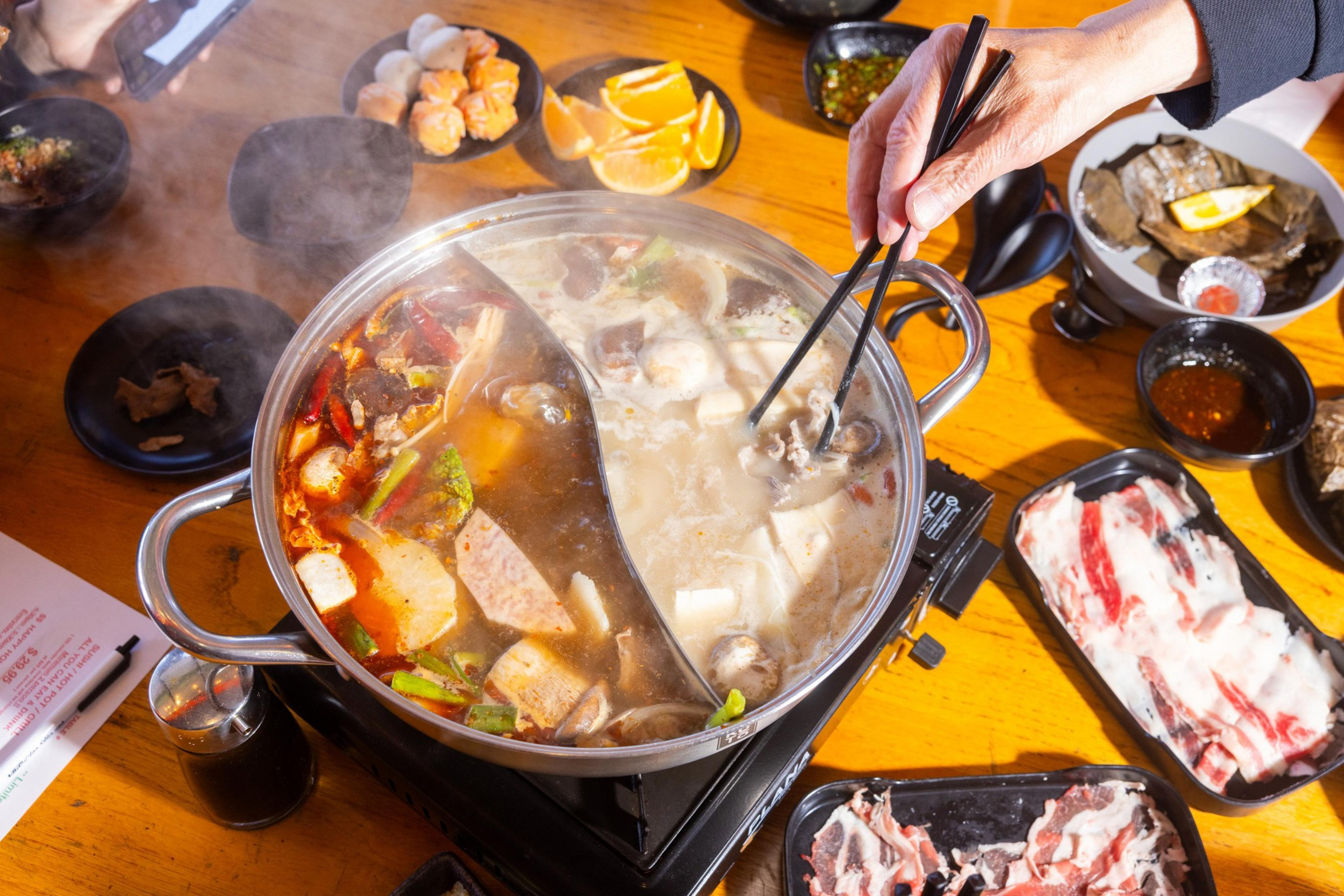 A divided hot pot with spicy and mild broths sits steaming, surrounded by plates of sliced meat, vegetables, orange slices, and dipping sauces. A hand uses chopsticks.