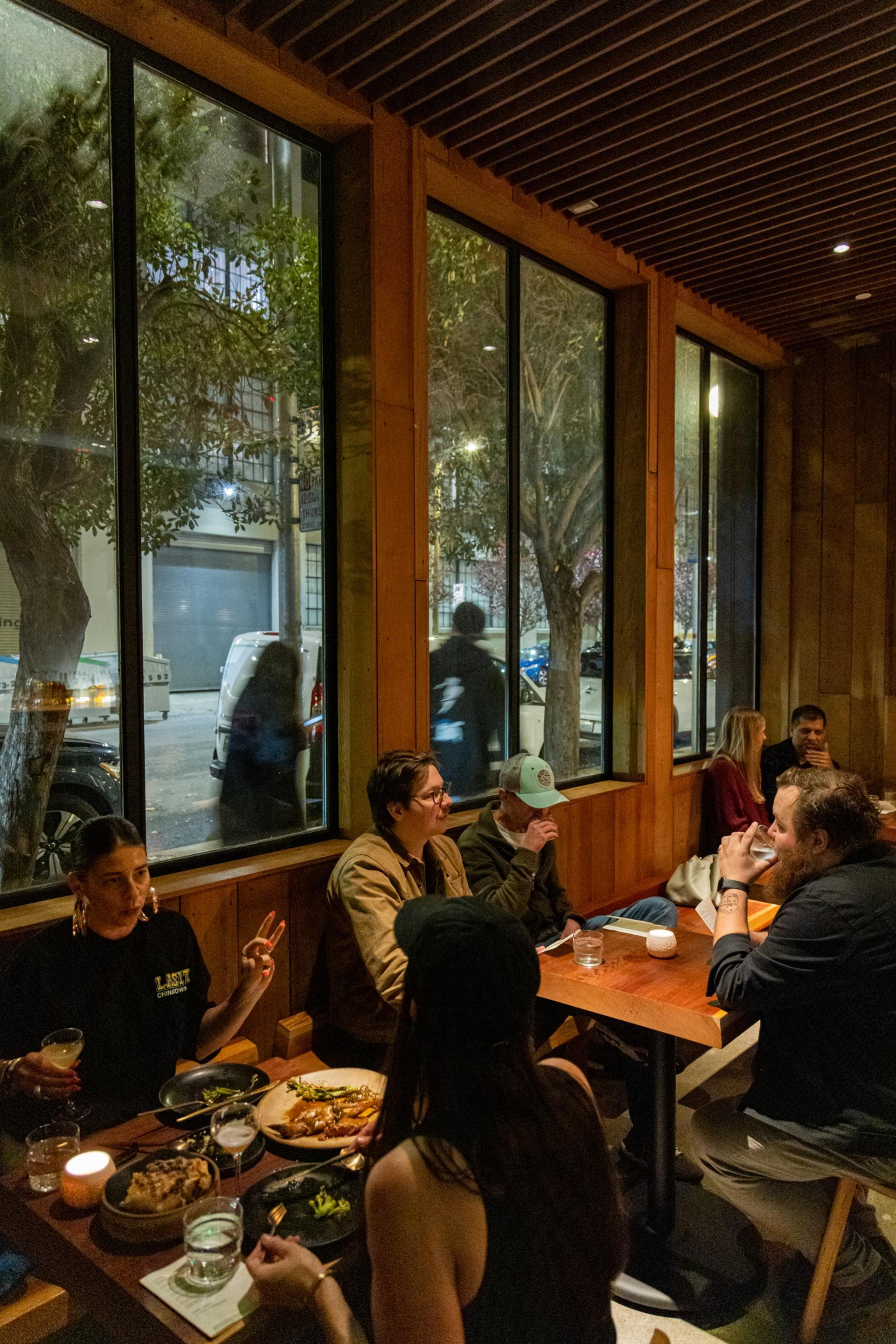 People are dining in a cozy restaurant with wooden interiors and large windows. They're chatting and enjoying their food, while the outside view shows a street.