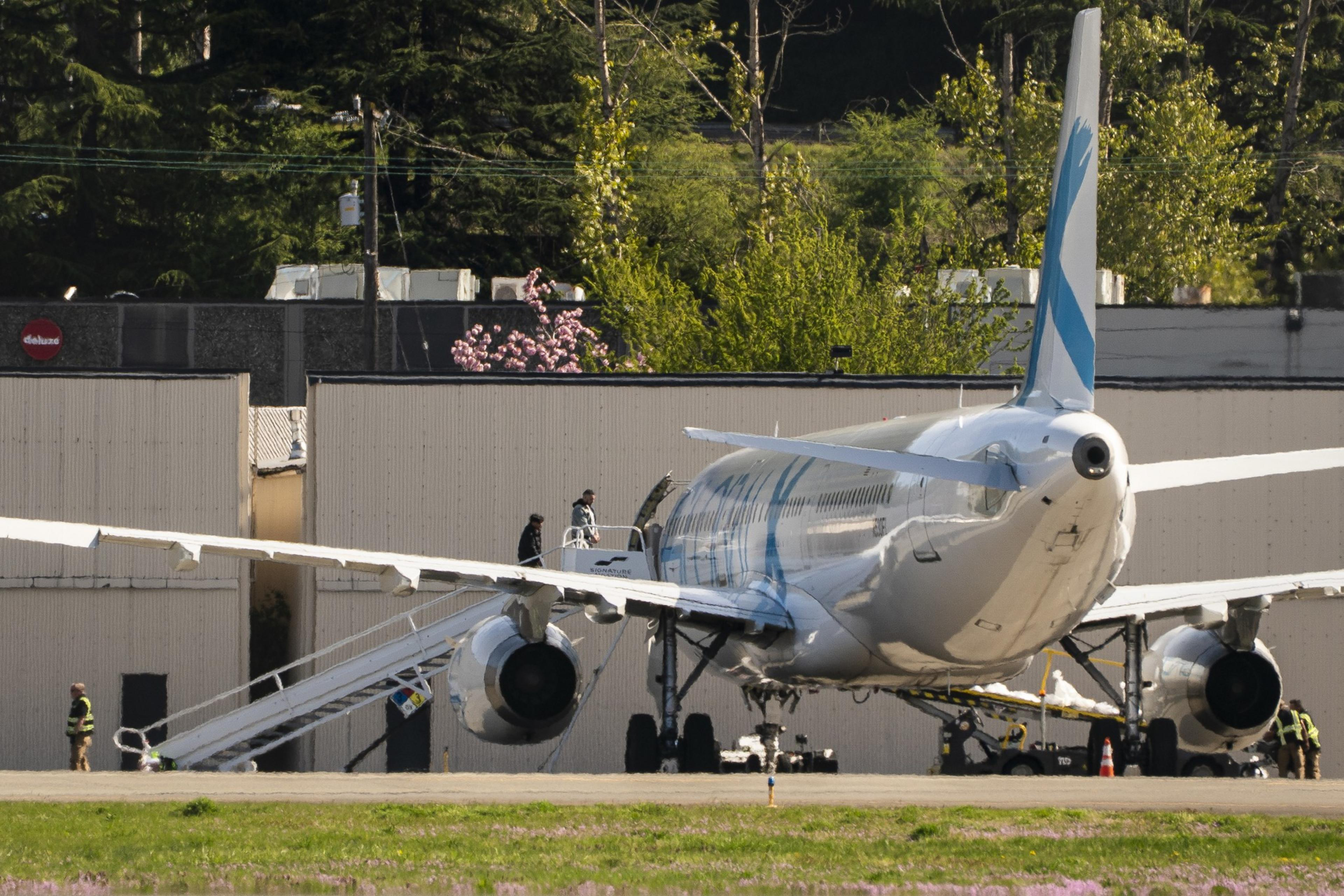 A white airplane is parked with its tail toward the viewer. Several people are near the rear with stairs attached, and there are trees and buildings in the background.