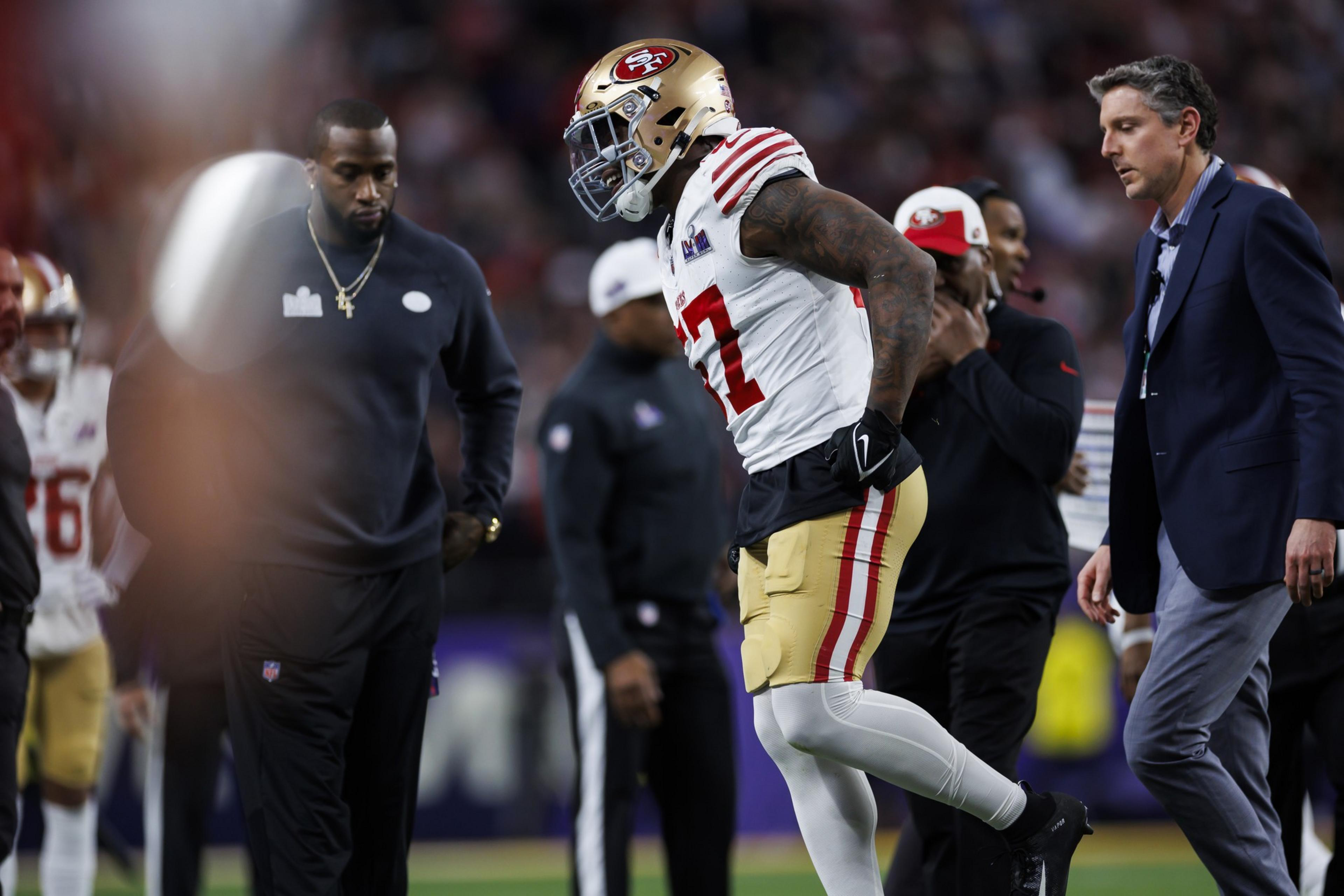 A football player in a 49ers uniform looks distressed, surrounded by two men in casual and formal attire, on a football field with blurred onlookers.