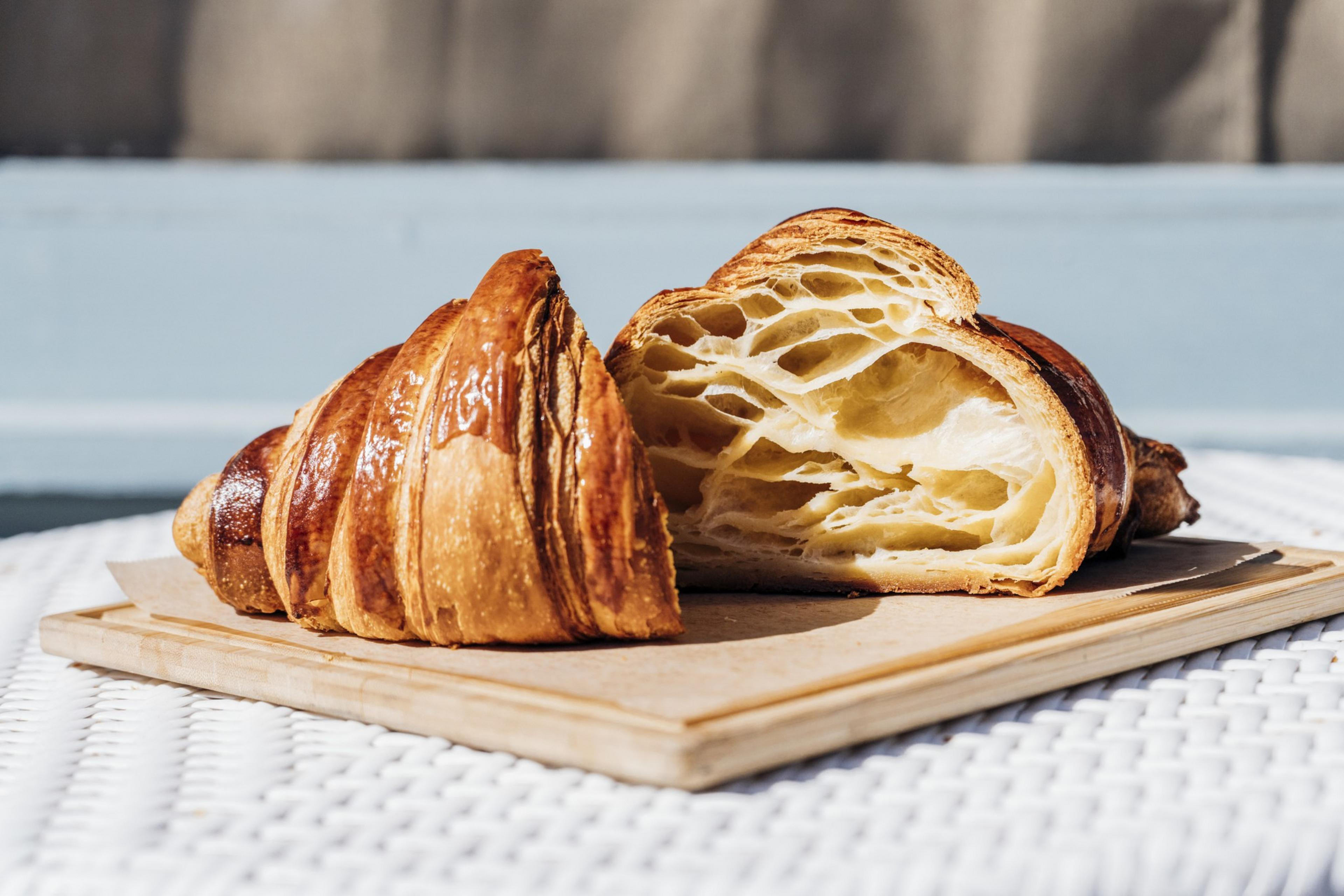 A croissant is cut in half, showing its flaky, golden-brown exterior and airy, layered interior, placed on a wooden board.