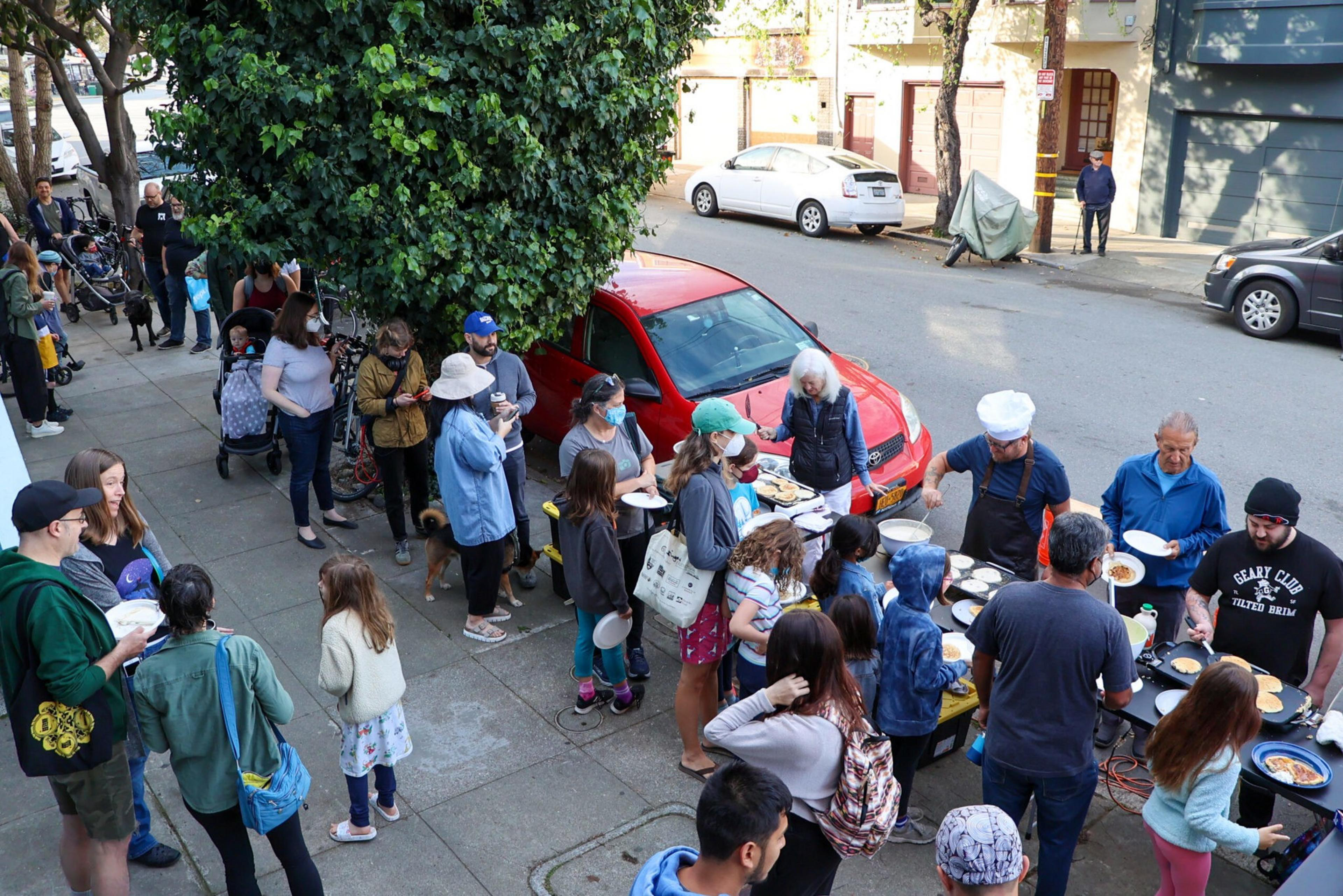A group of people, including adults and children, gather on a sidewalk for a pancake event. Some are in line, while others serve pancakes from a table.