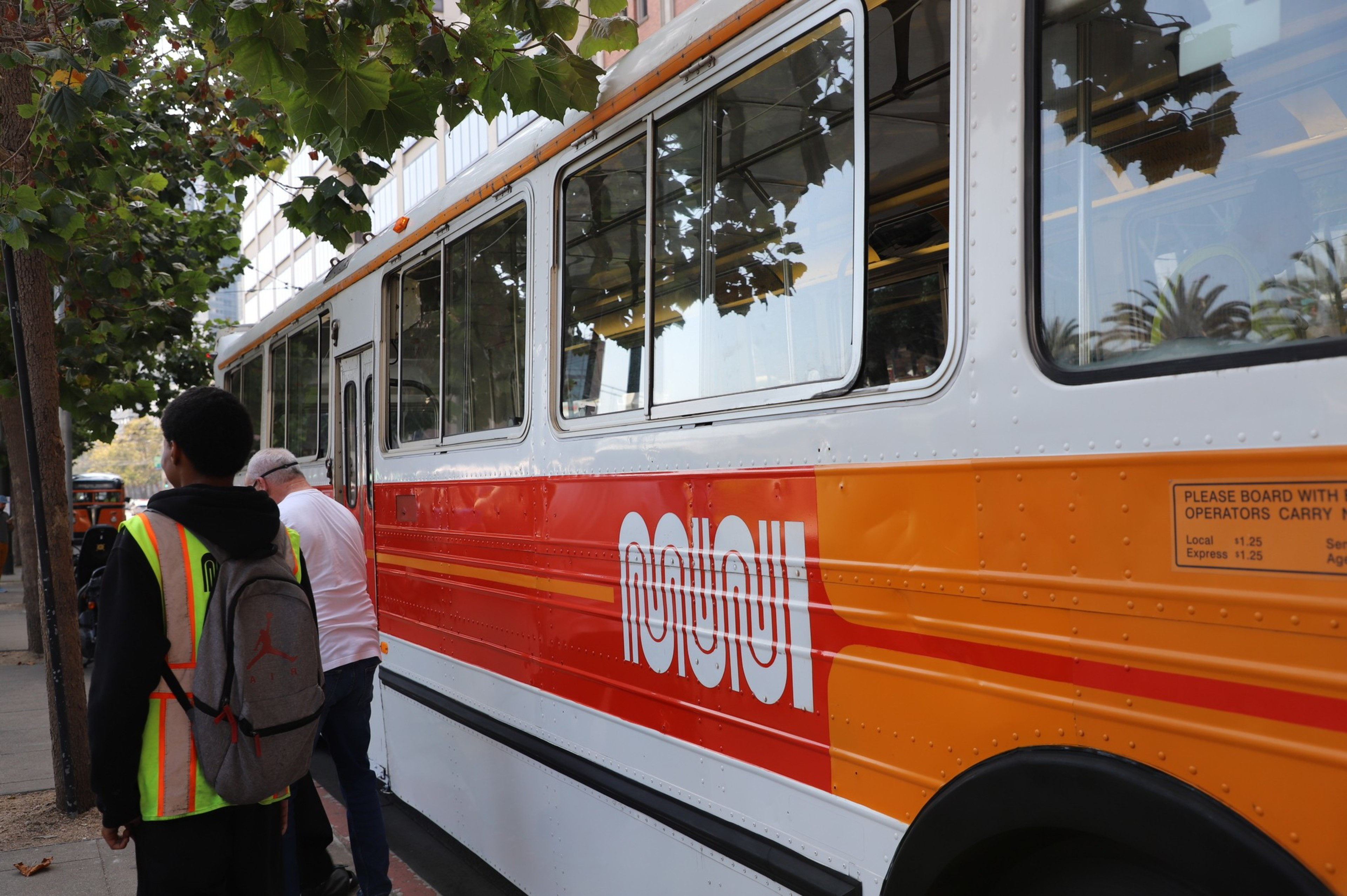 A person in a high-visibility vest is boarding an orange and white city bus on a tree-lined street.
