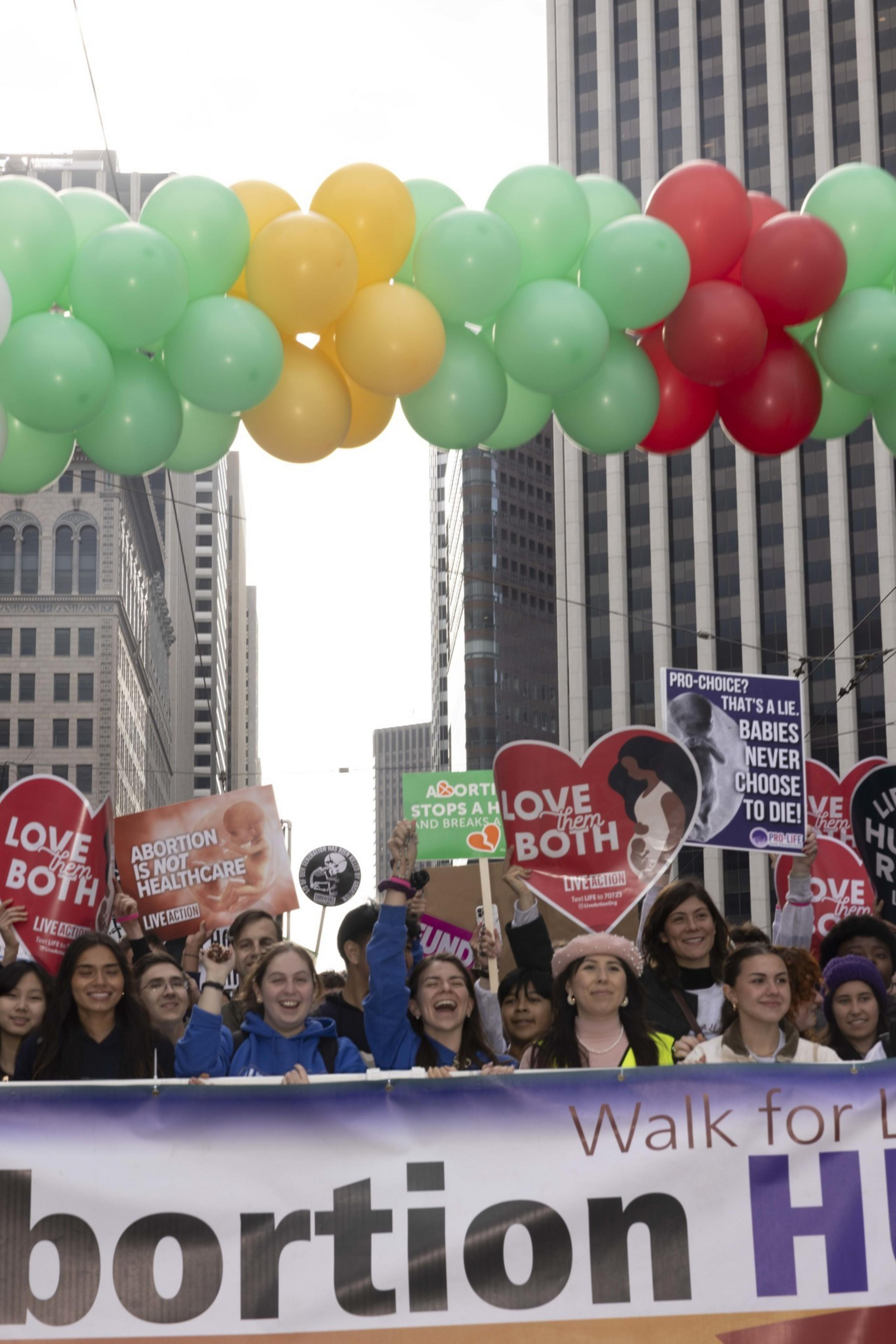 A group of people in an urban setting hold signs with anti-abortion messages. Colorful balloons form an arch overhead, and buildings are visible in the background.