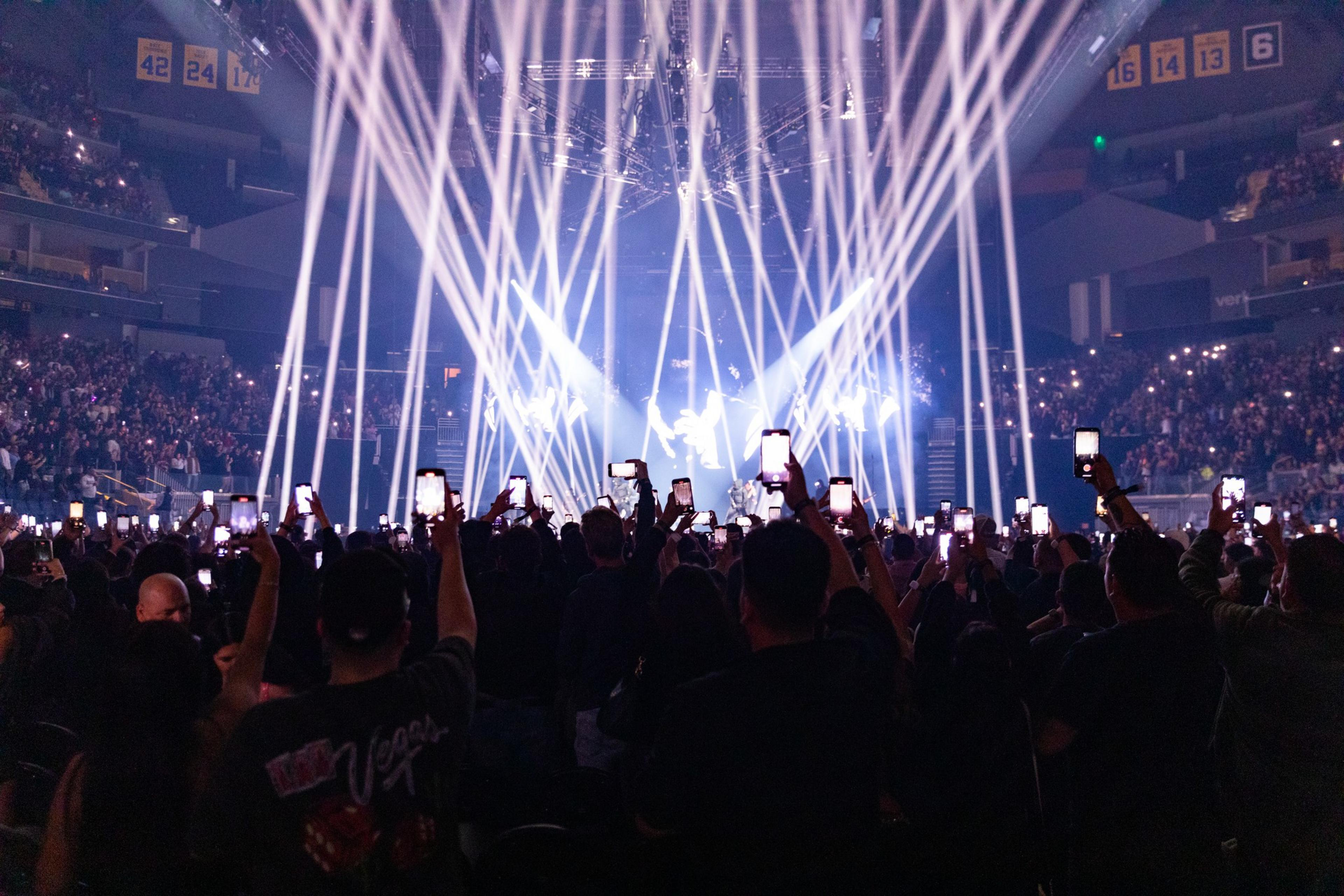 A large concert with bright stage lights beaming into the audience. Many attendees are holding up smartphones to record or photograph the performance.