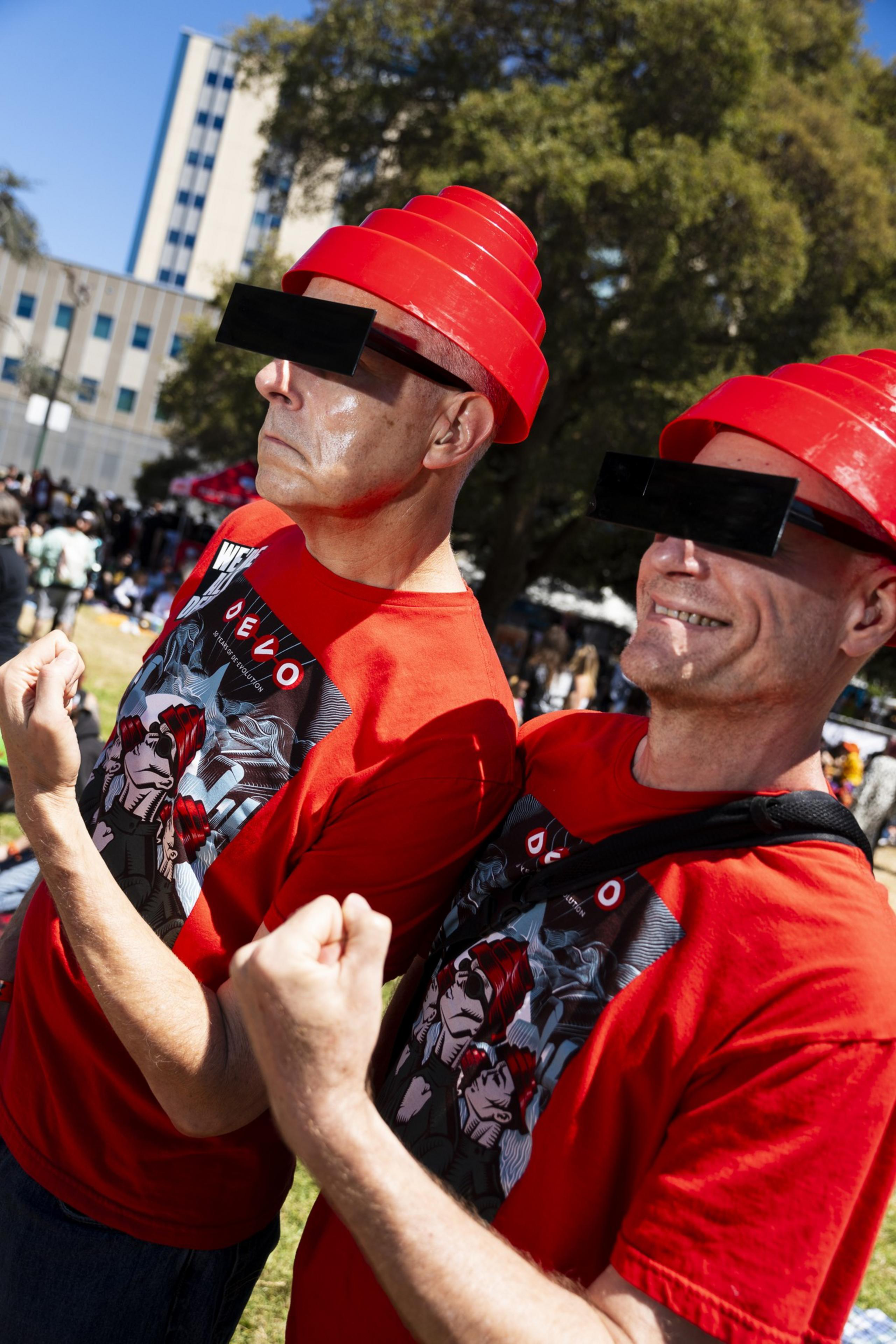 Two people in red hats and sunglasses, wearing matching red shirts with graphic designs, pose outdoors in a lively setting with trees and buildings.