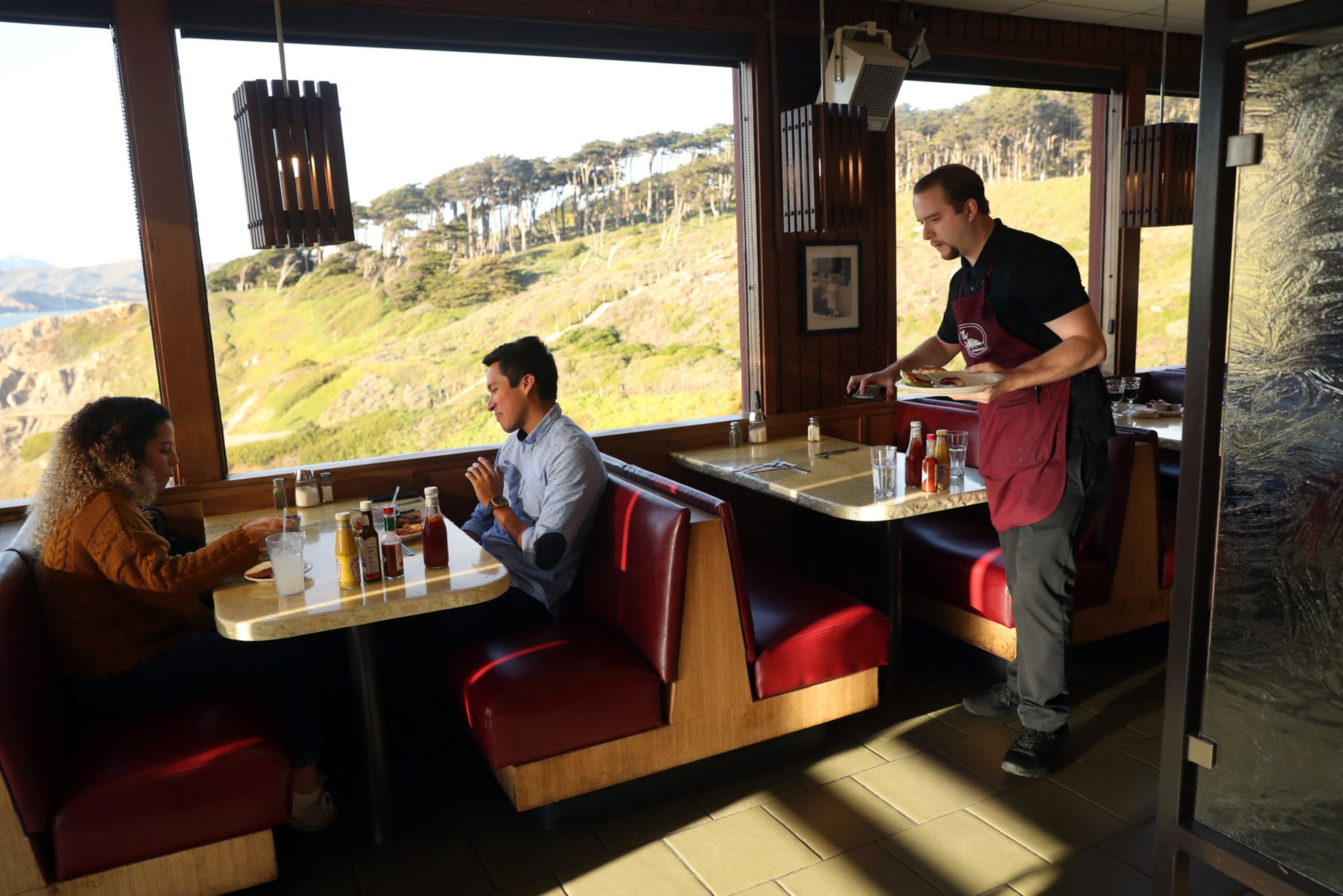 Two people sit at a diner booth eating, while a waiter wearing a red apron serves food at another booth, with a scenic hillside visible outside.