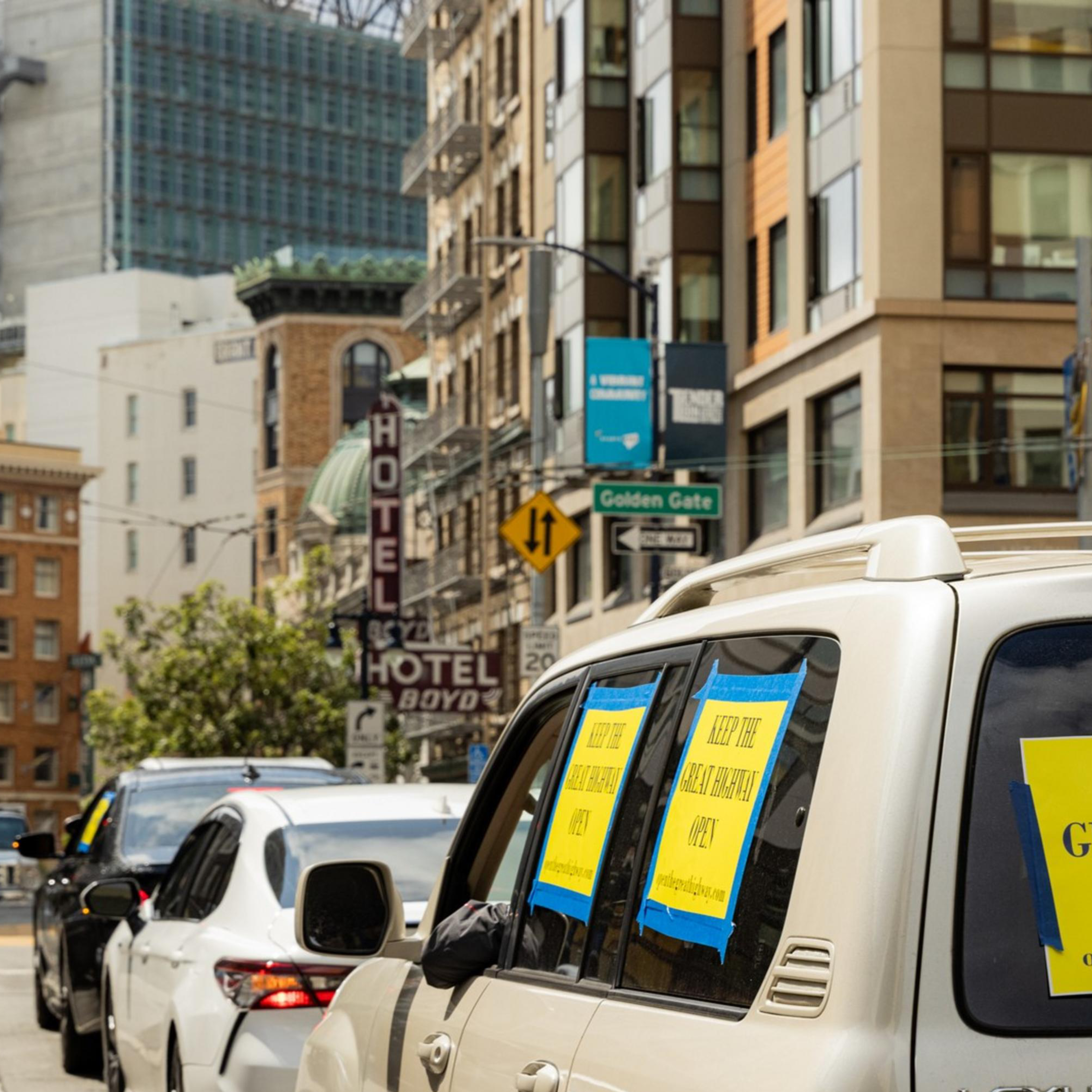 In a city with tall buildings, cars are lined up on the street. The leading car, a Lexus, has yellow signs saying &quot;KEEP THE GREAT HIGHWAY OPEN&quot; taped to its windows.