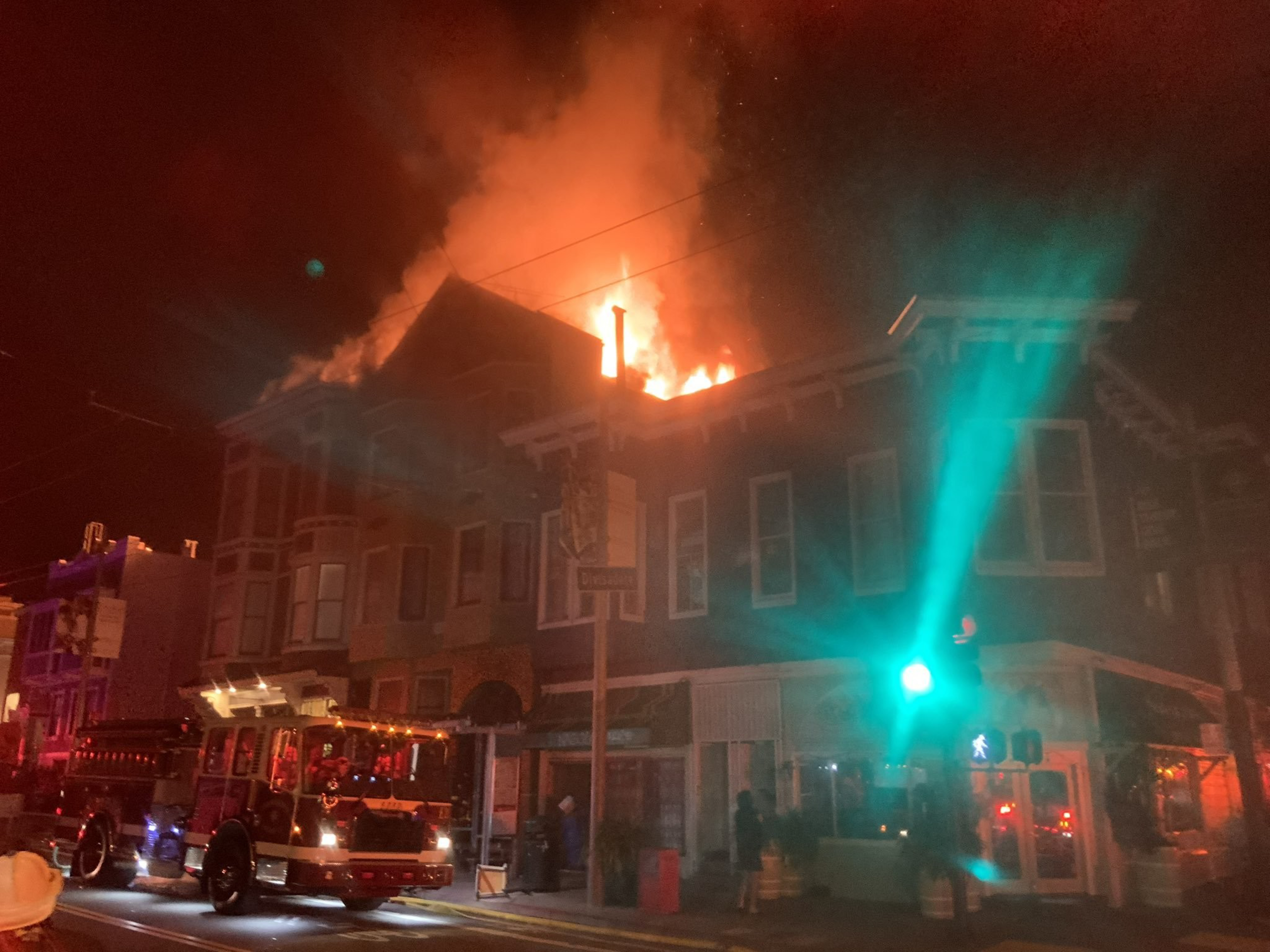 A building's roof is engulfed in flames at night, with a fire truck parked nearby and a green traffic light illuminating the scene.