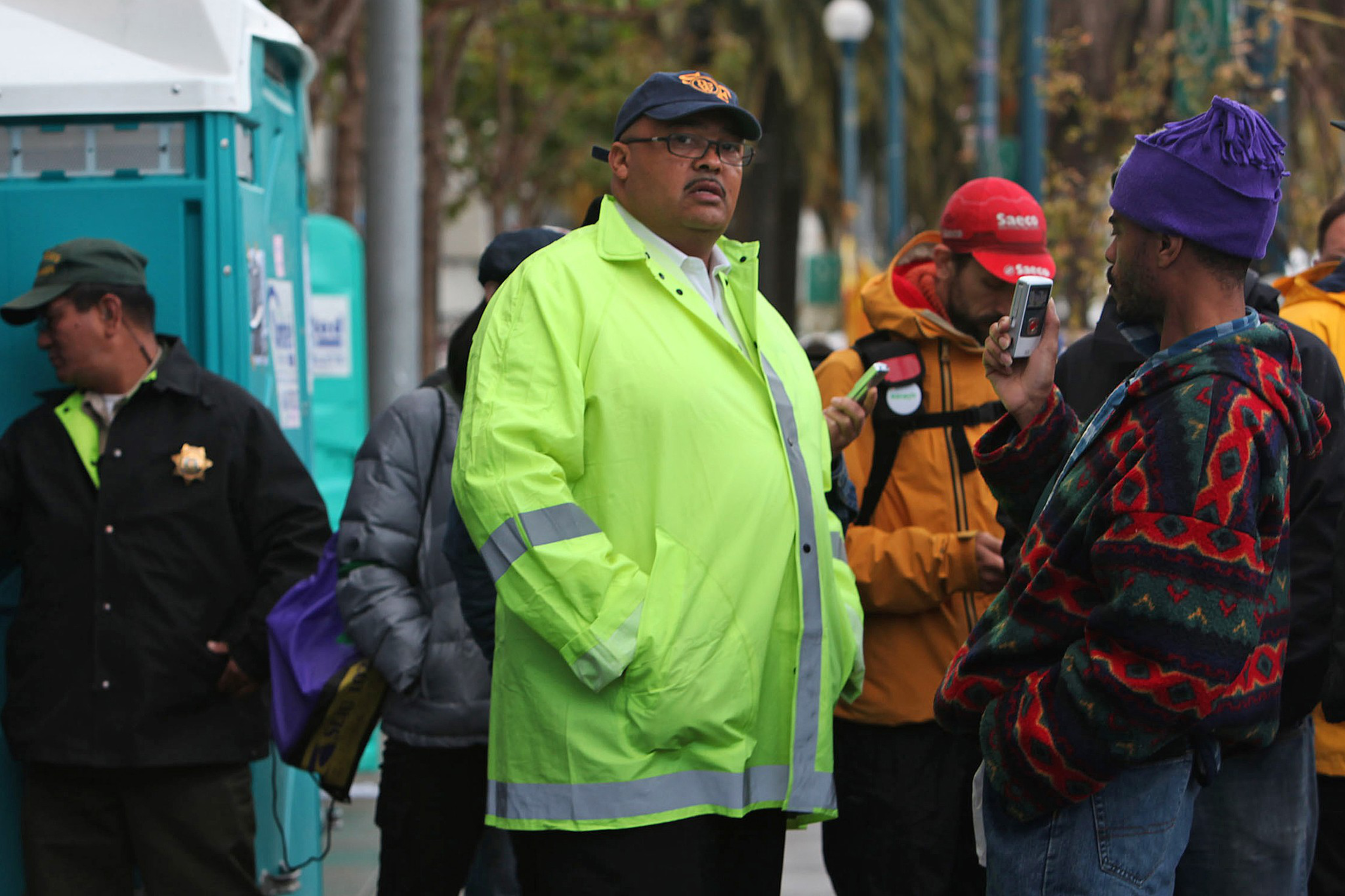 A man in a bright green jacket stands in a city plaza with other people around.