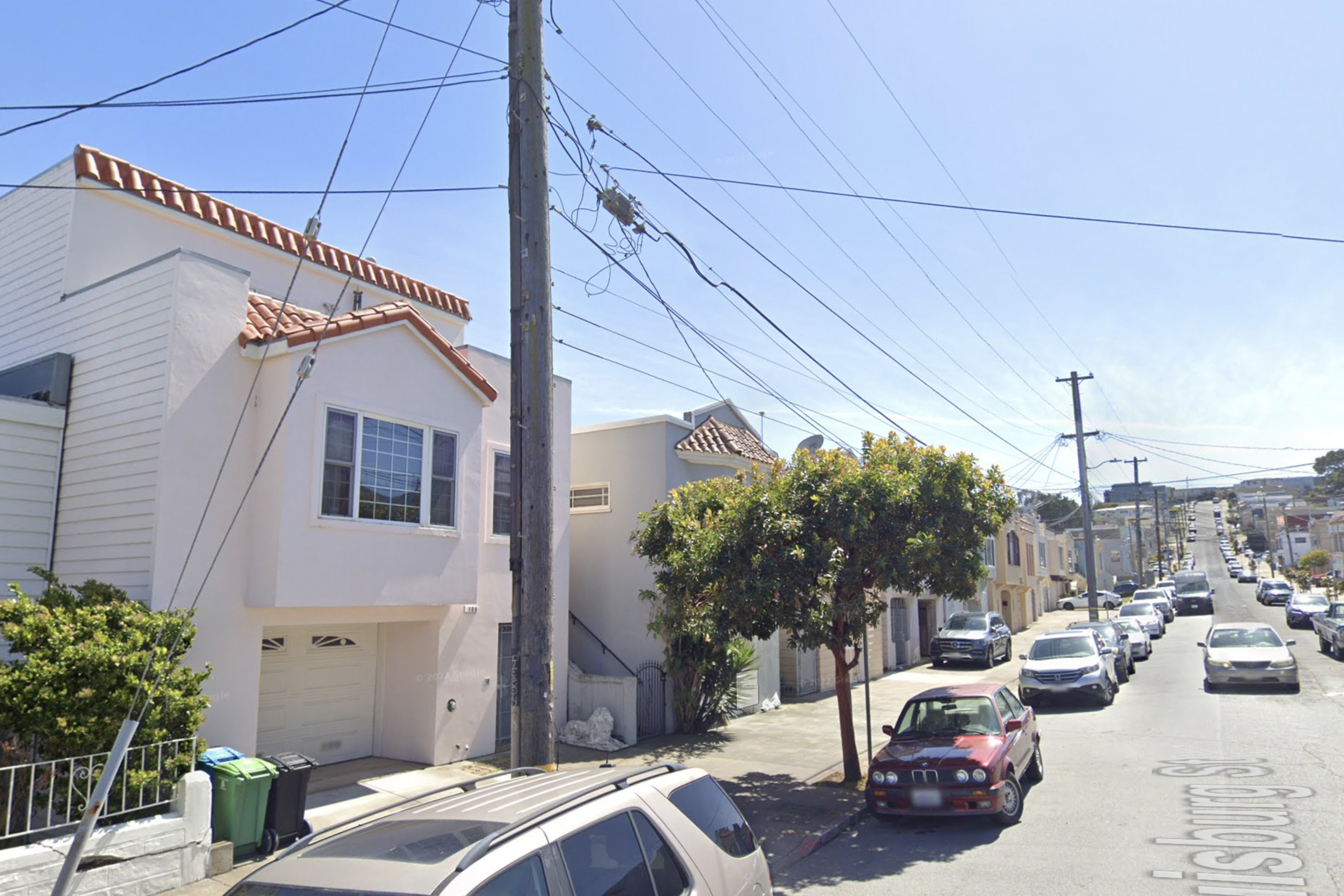 A residential street lined with houses, parked cars, a tree on the sidewalk, and power lines overhead stretching into the distance.