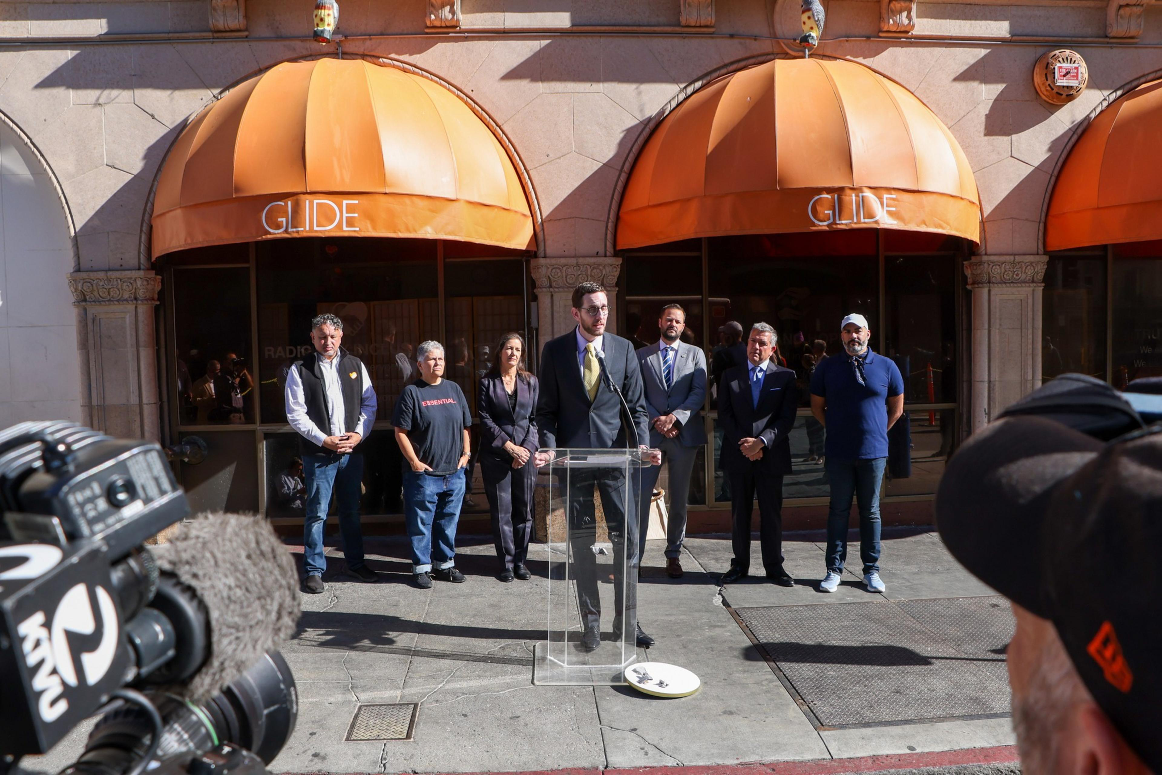 A man in a suit speaks at a clear podium outside a building with orange awnings labeled "GLIDE," flanked by six people and filmed by cameras.