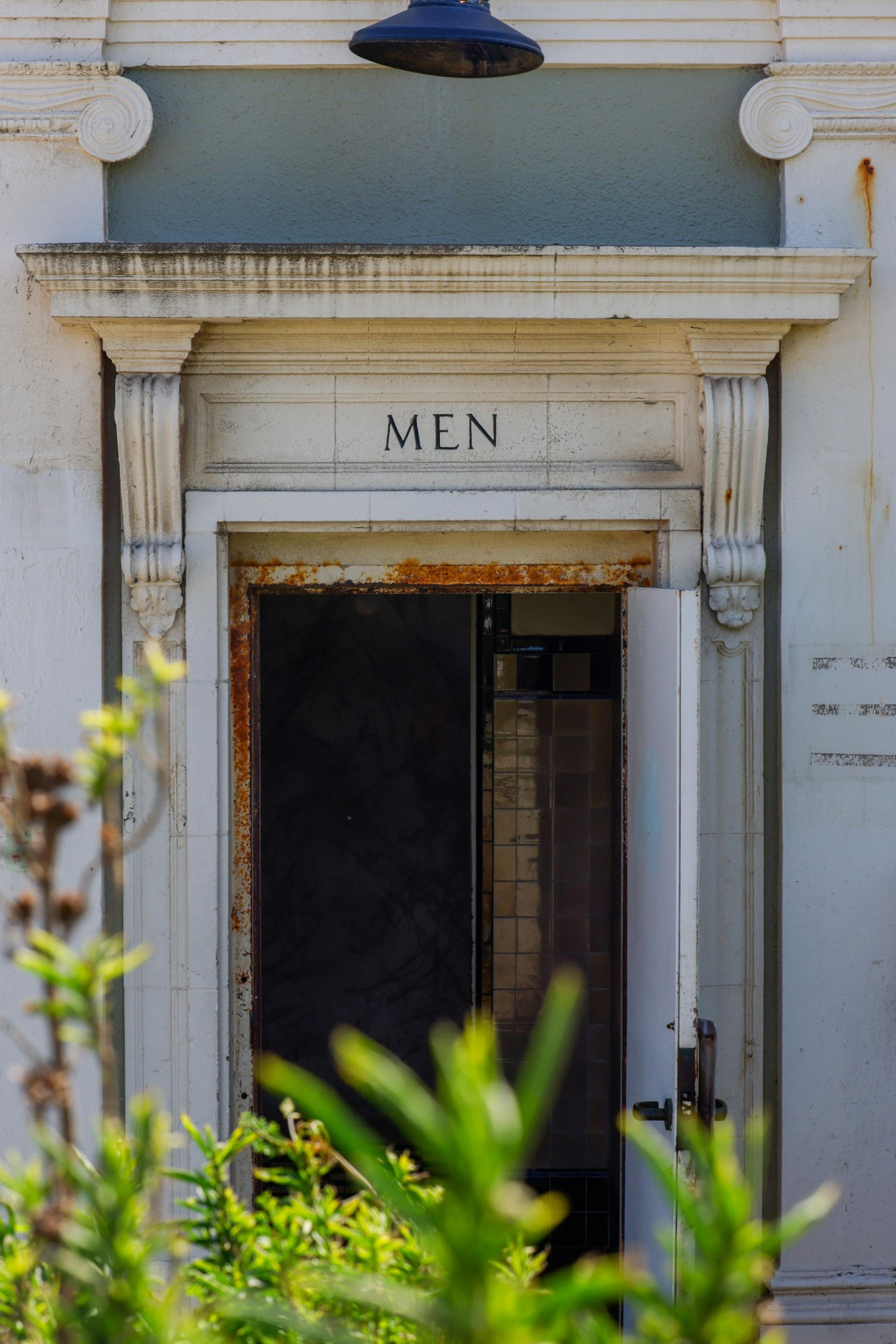An open men's restroom door, framed by an ornate, slightly weathered archway with columns. The scene includes overgrown plants in the foreground.