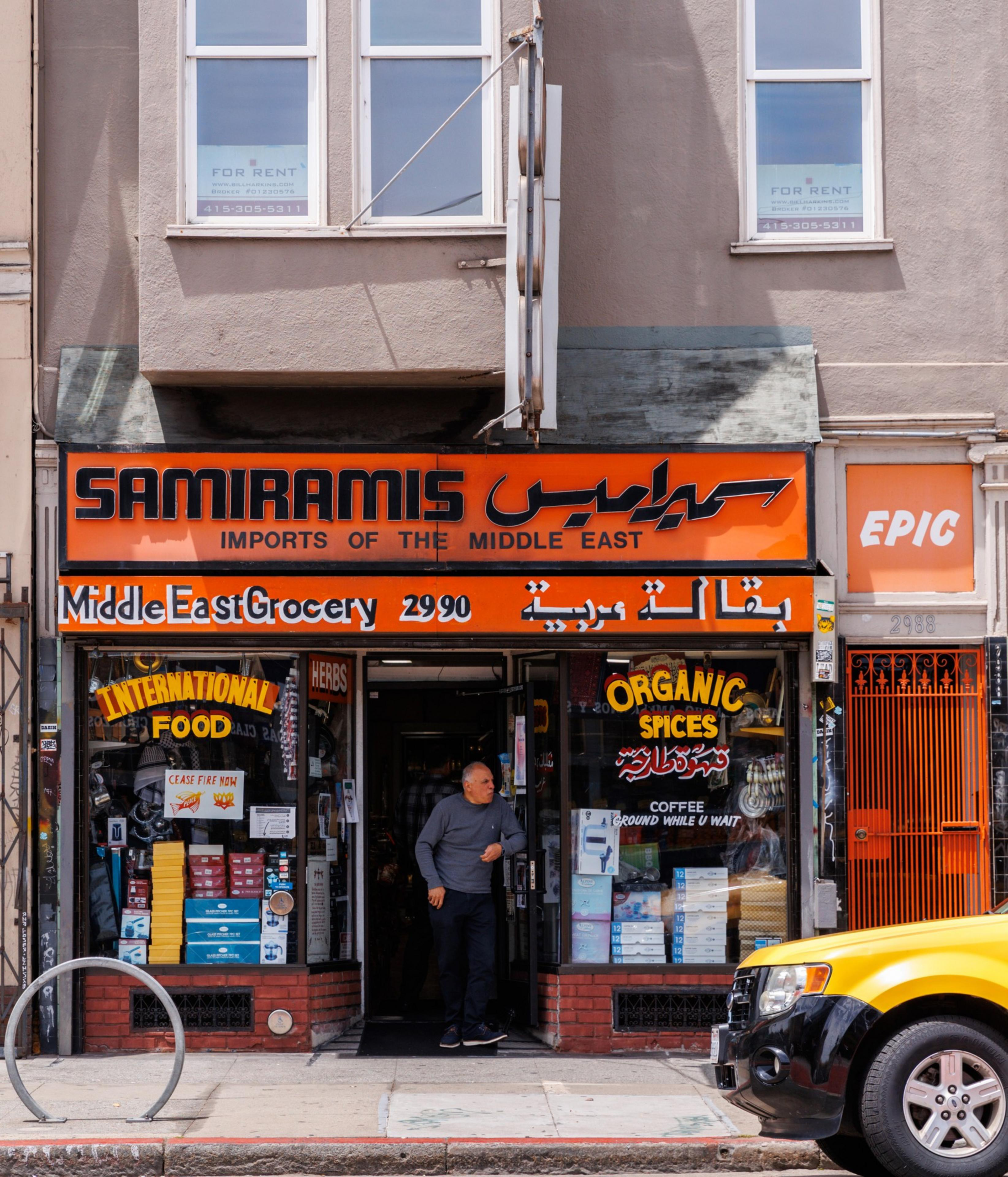 A man stands at the entrance of &quot;Samiramis Middle East Grocery,&quot; an orange storefront with signs advertising organic spices, international food, and products inside.