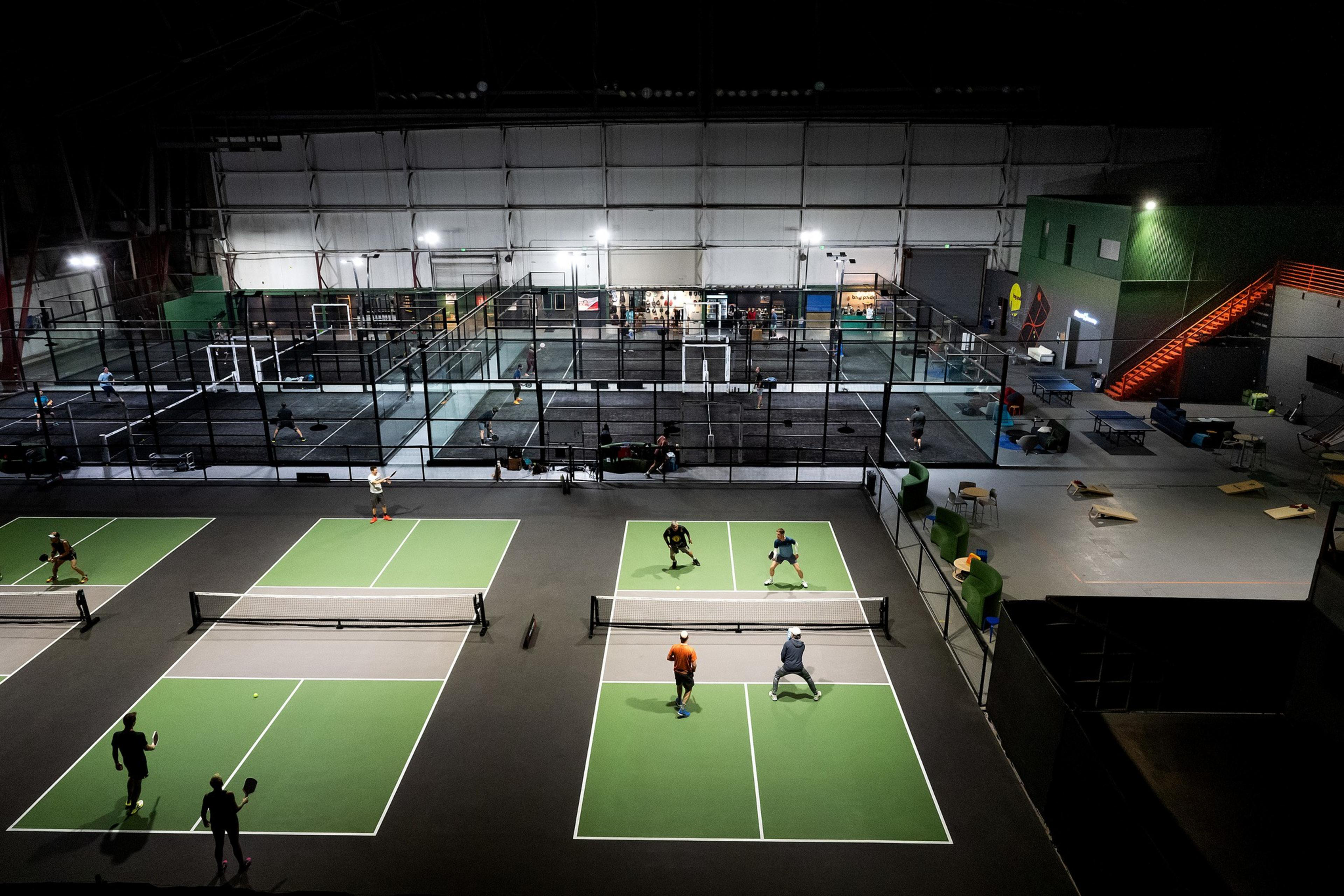 The image shows multiple people playing indoor pickleball on green courts with nets. It's a large sports facility with seating and a staircase visible nearby.