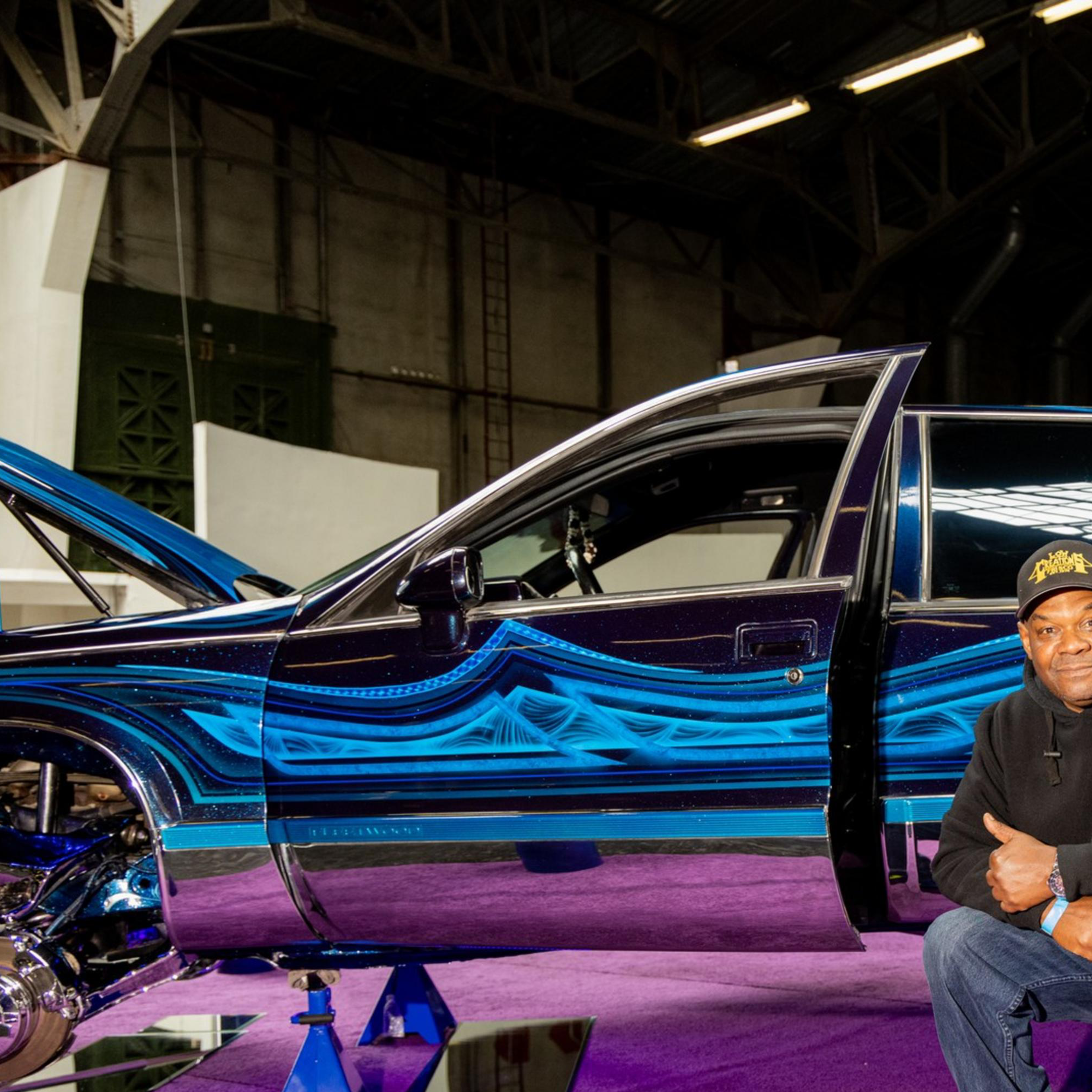 A man kneels on a purple carpet beside a custom car with intricate blue patterns and opened hood. The car is lifted on stands, showcasing chrome details.