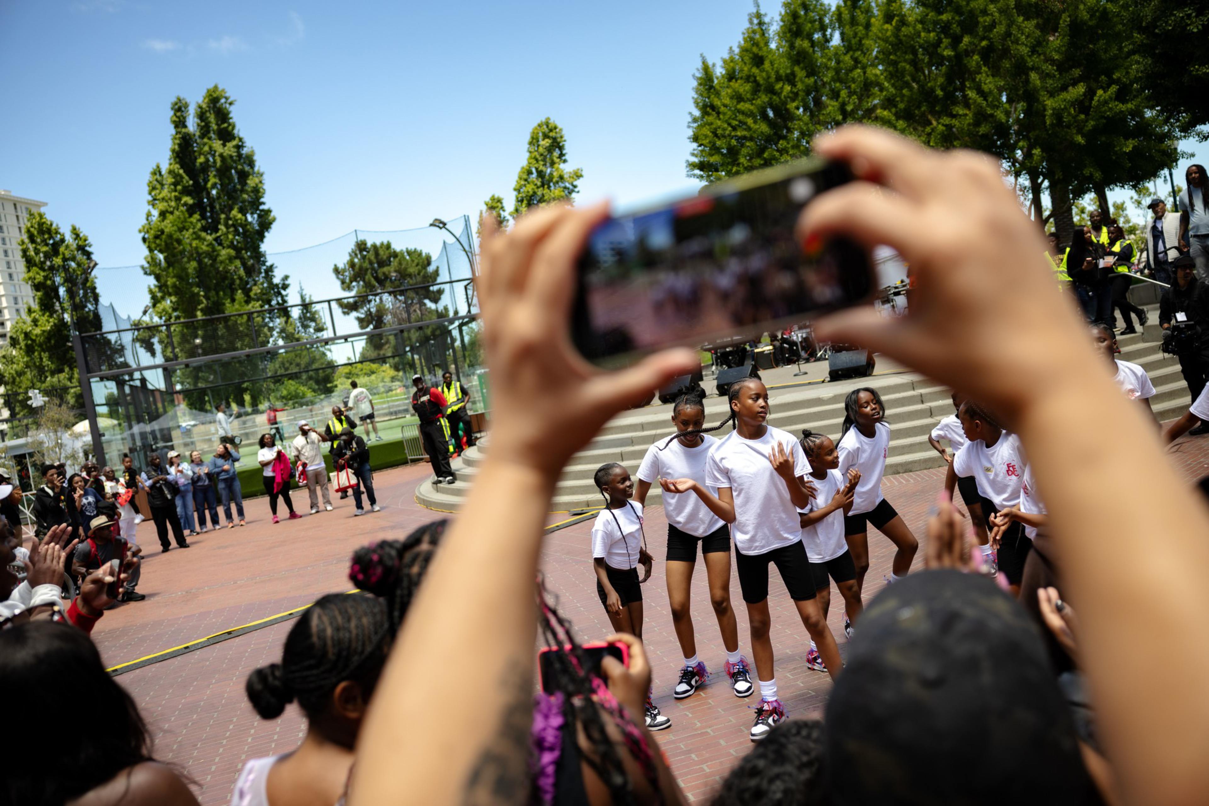 A group of young people in white shirts and black shorts dance in an outdoor plaza while spectators watch and record them on phones.