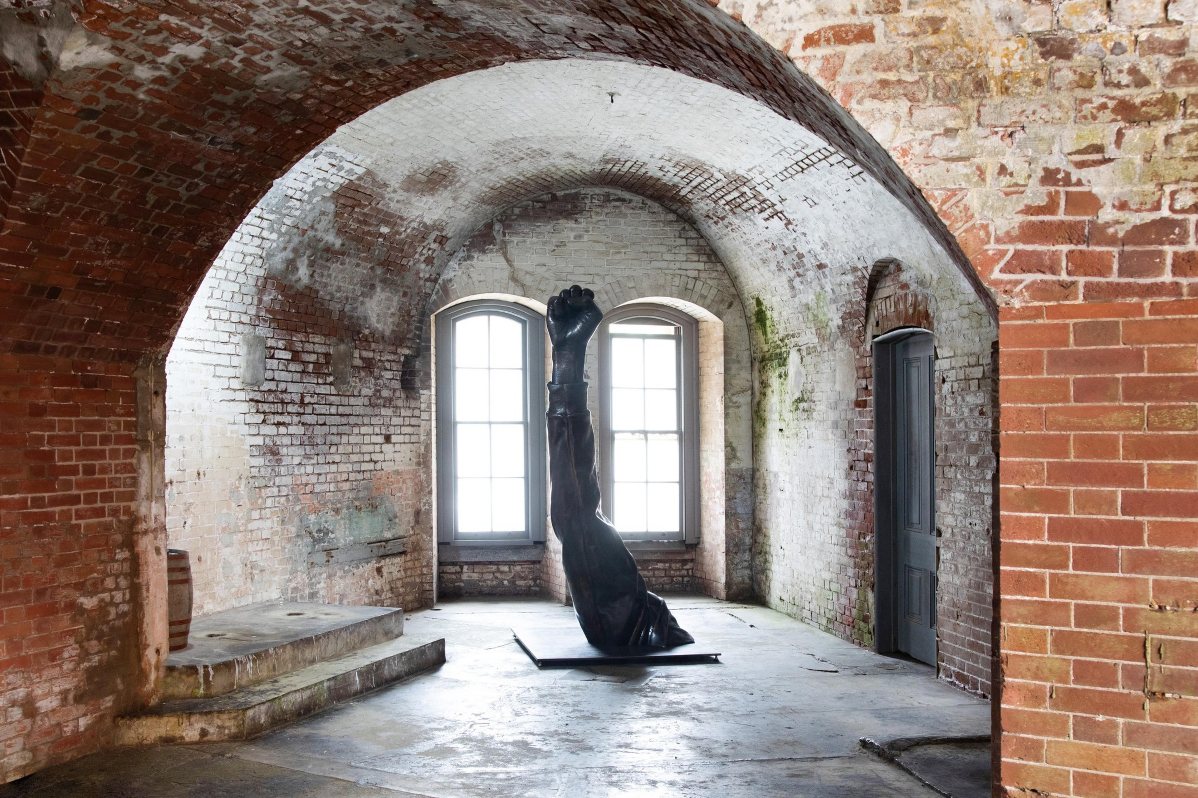 A brick-walled room with arched ceilings features a sculpture of an arm with a clenched fist emerging from the floor, set against two large, arched windows.