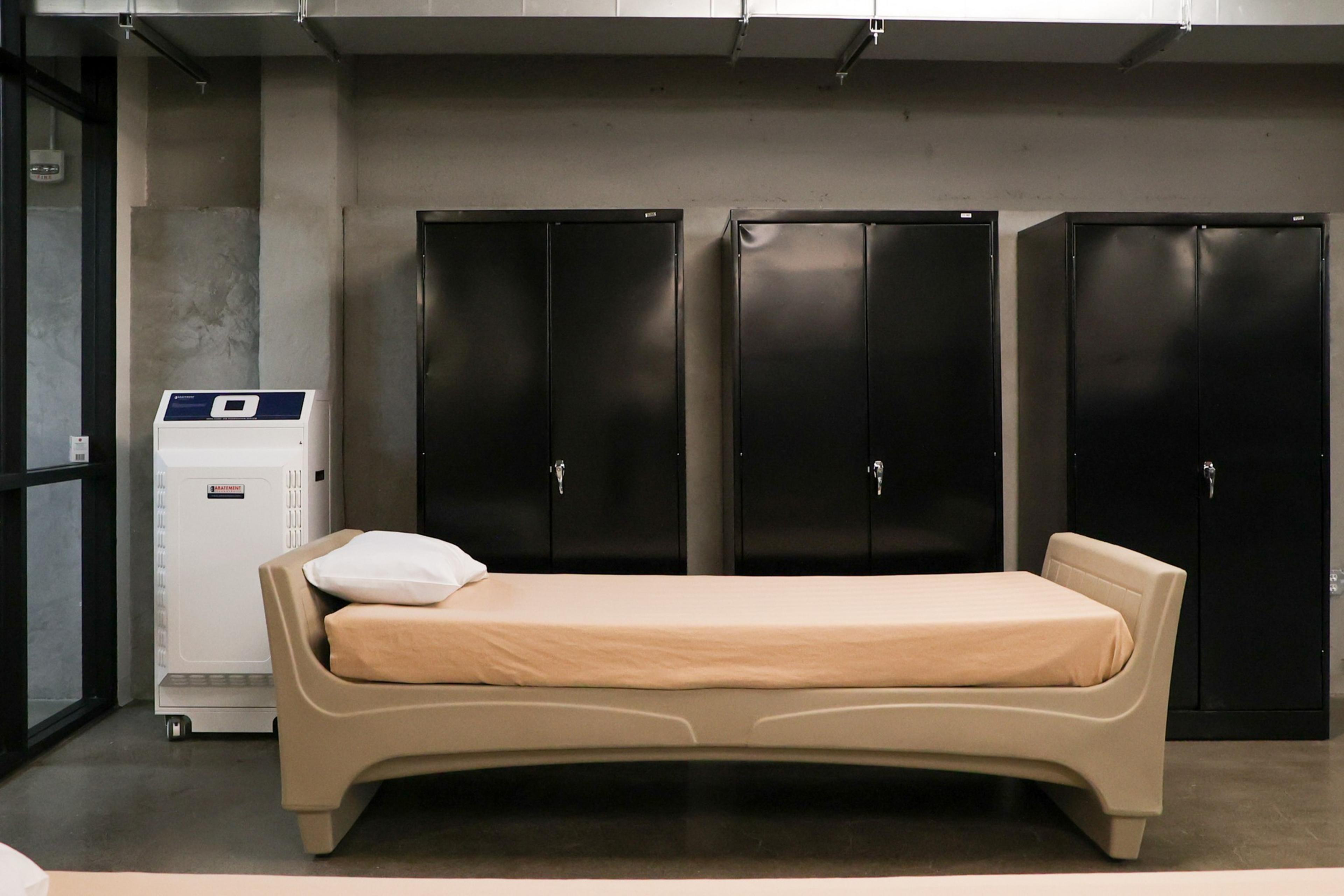 A simple beige bed with a pillow and a folded blanket is placed in front of three closed black metal lockers and a white machine on a polished floor.