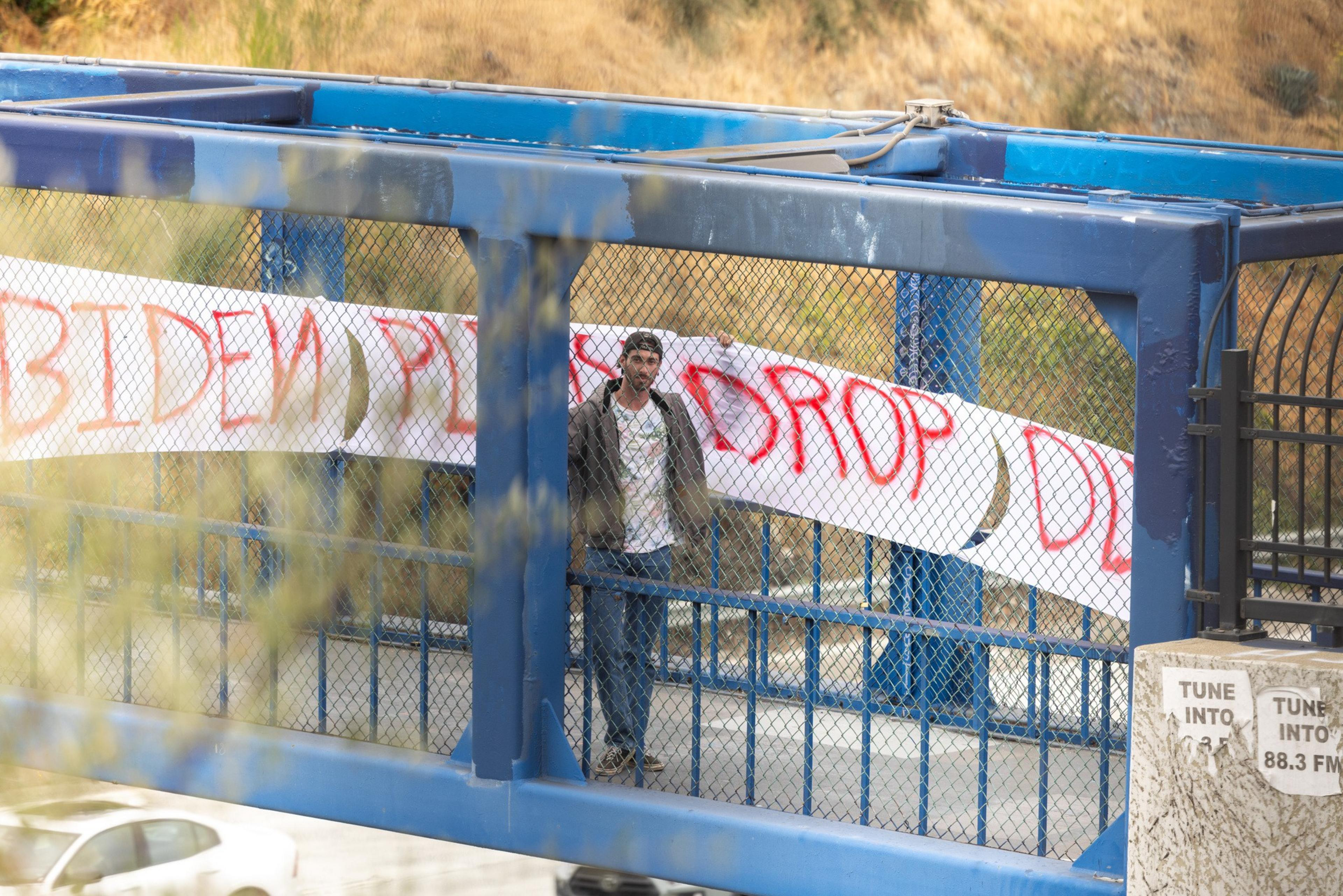 A person stands on a blue metal pedestrian bridge, holding a white banner with &quot;BIDEN PLEASE DROP&quot; written in red paint. A car is visible on the road below.