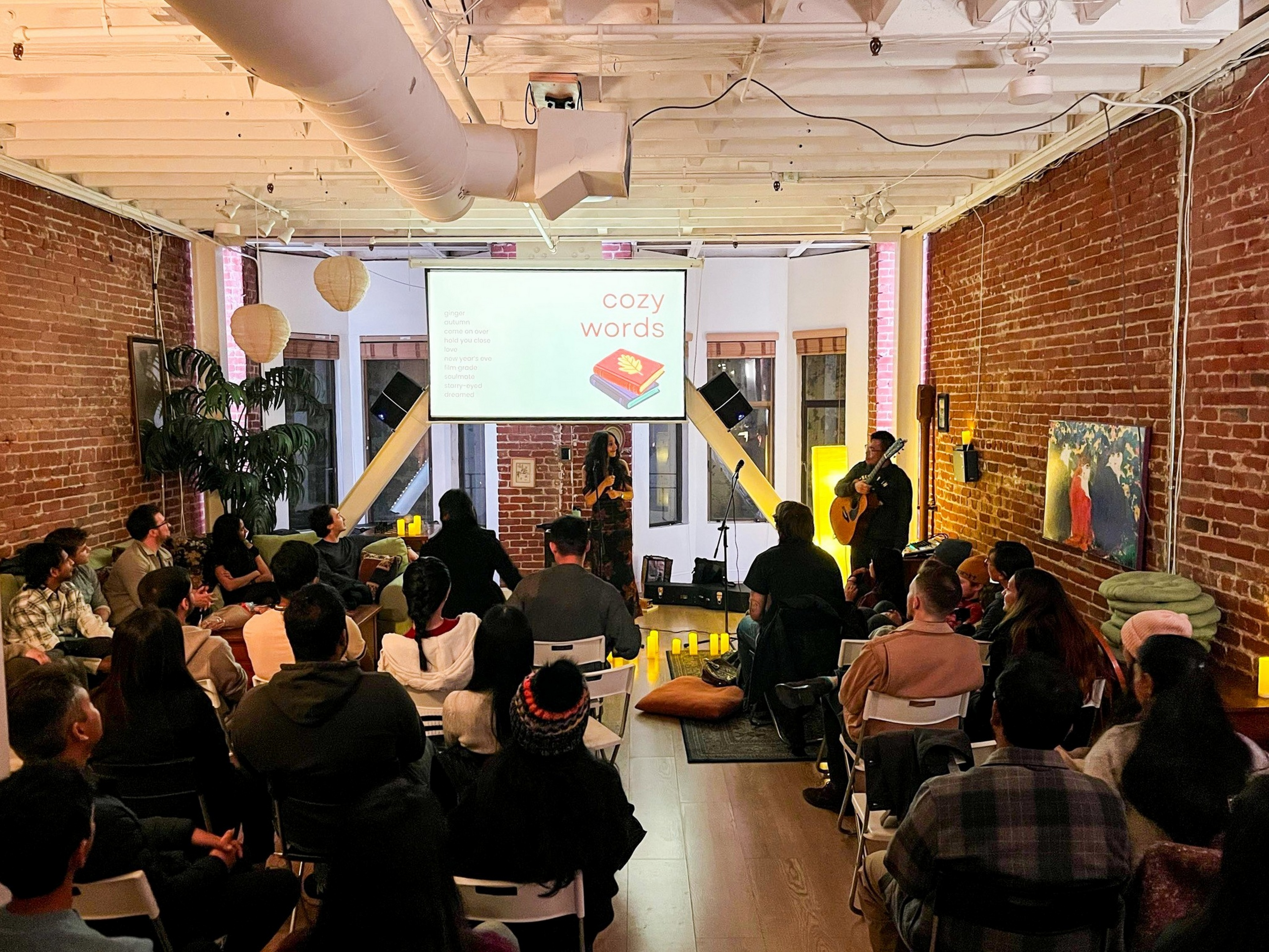 A group of people sit in a cozy room with exposed brick walls, listening to a woman speak and a man play guitar near a screen displaying “cozy words.”