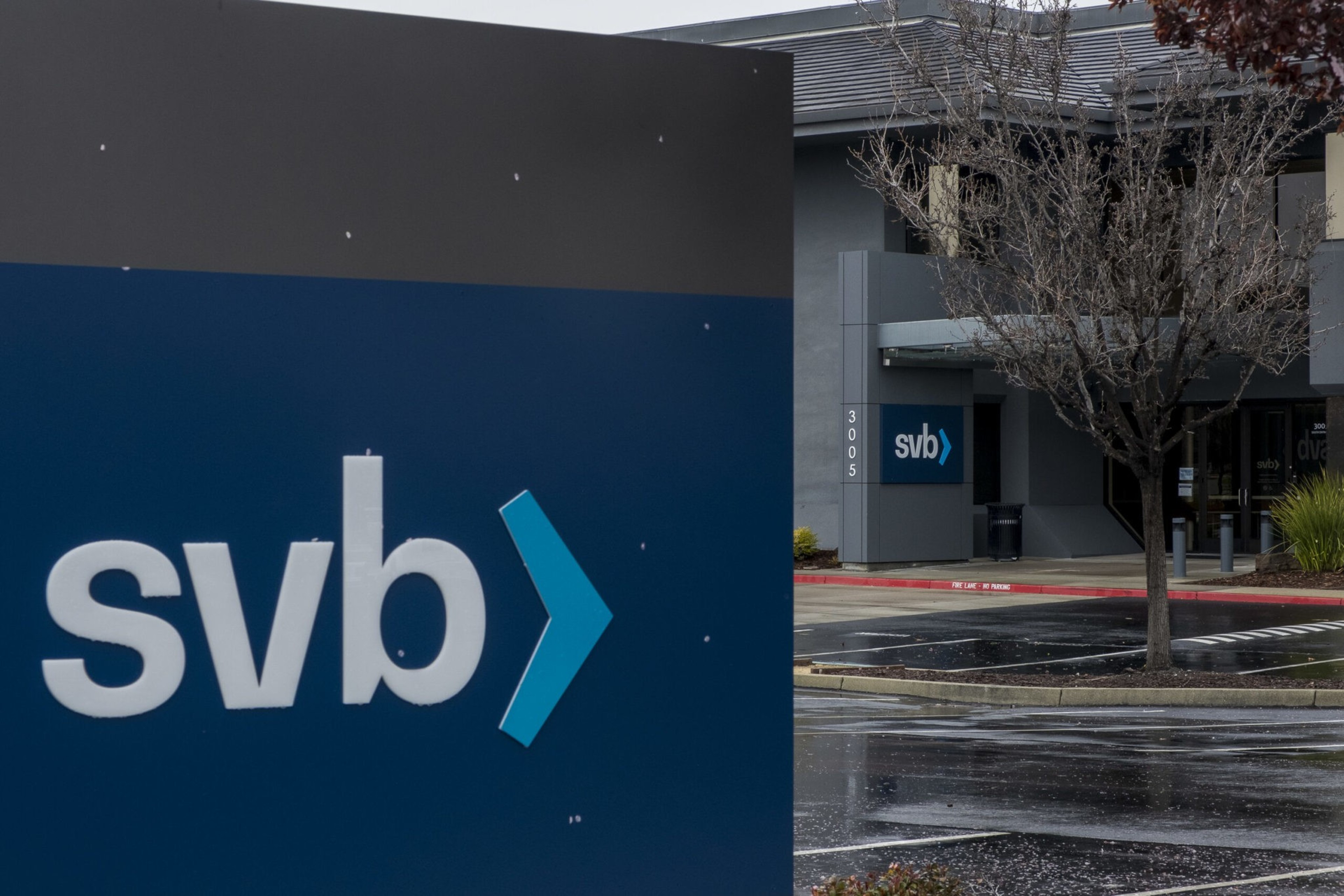 A large blue and gray sign with "svb" and a blue arrow stands outside a modern building with a smaller matching sign and a bare tree nearby.