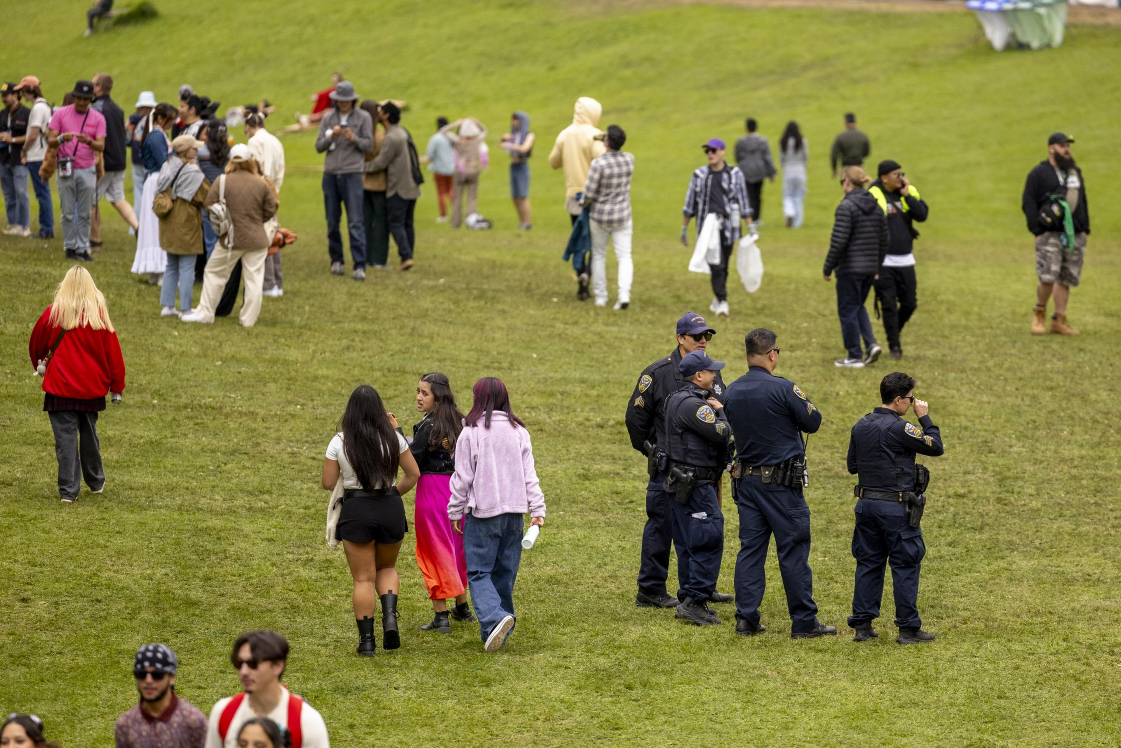 The image shows a grassy field with groups of people and several police officers. Some are walking, others are standing and chatting. The scene is casual and relaxed.