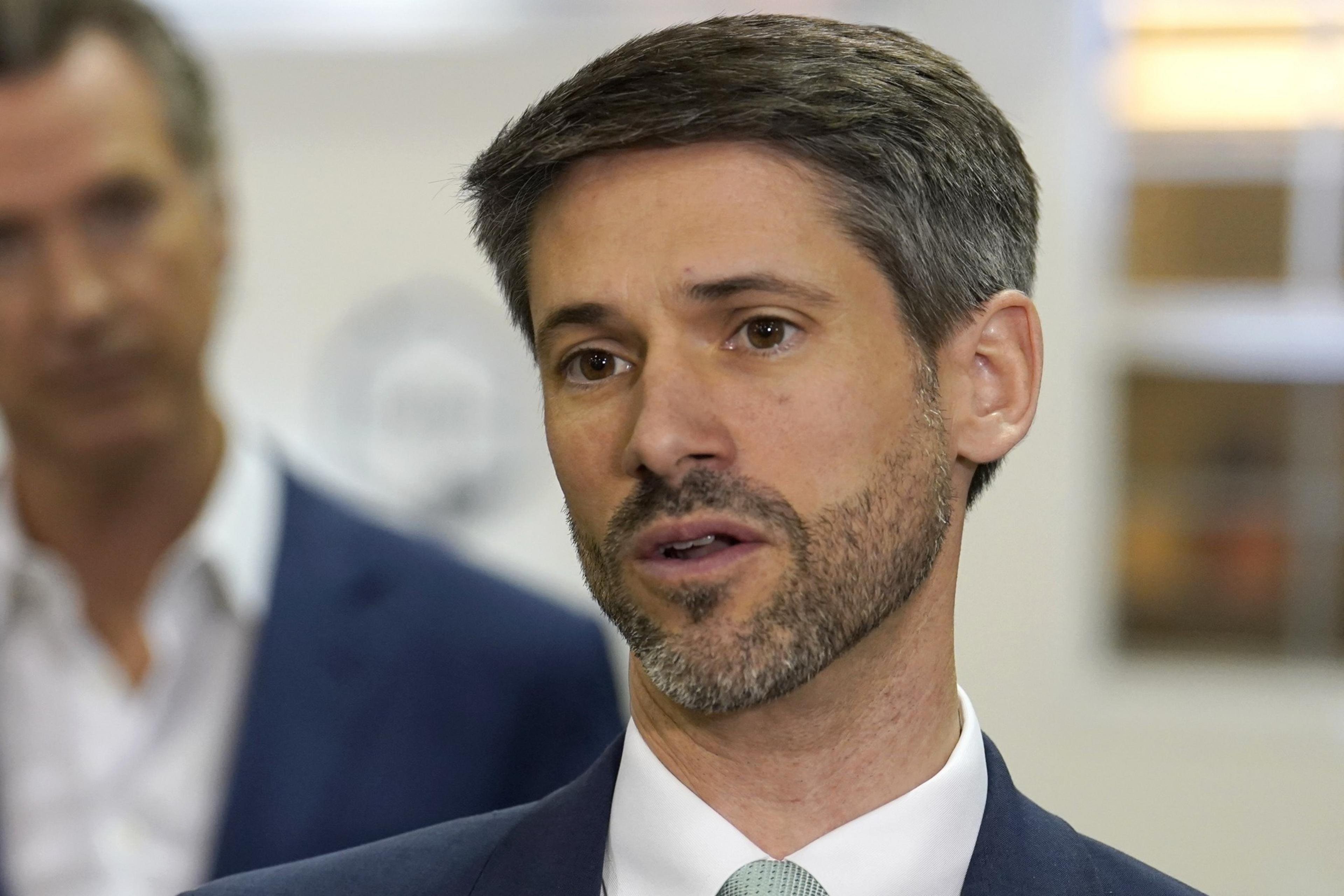 A man with short dark hair and a trimmed beard speaks while wearing a suit and tie, with another blurred person and a light background behind him.