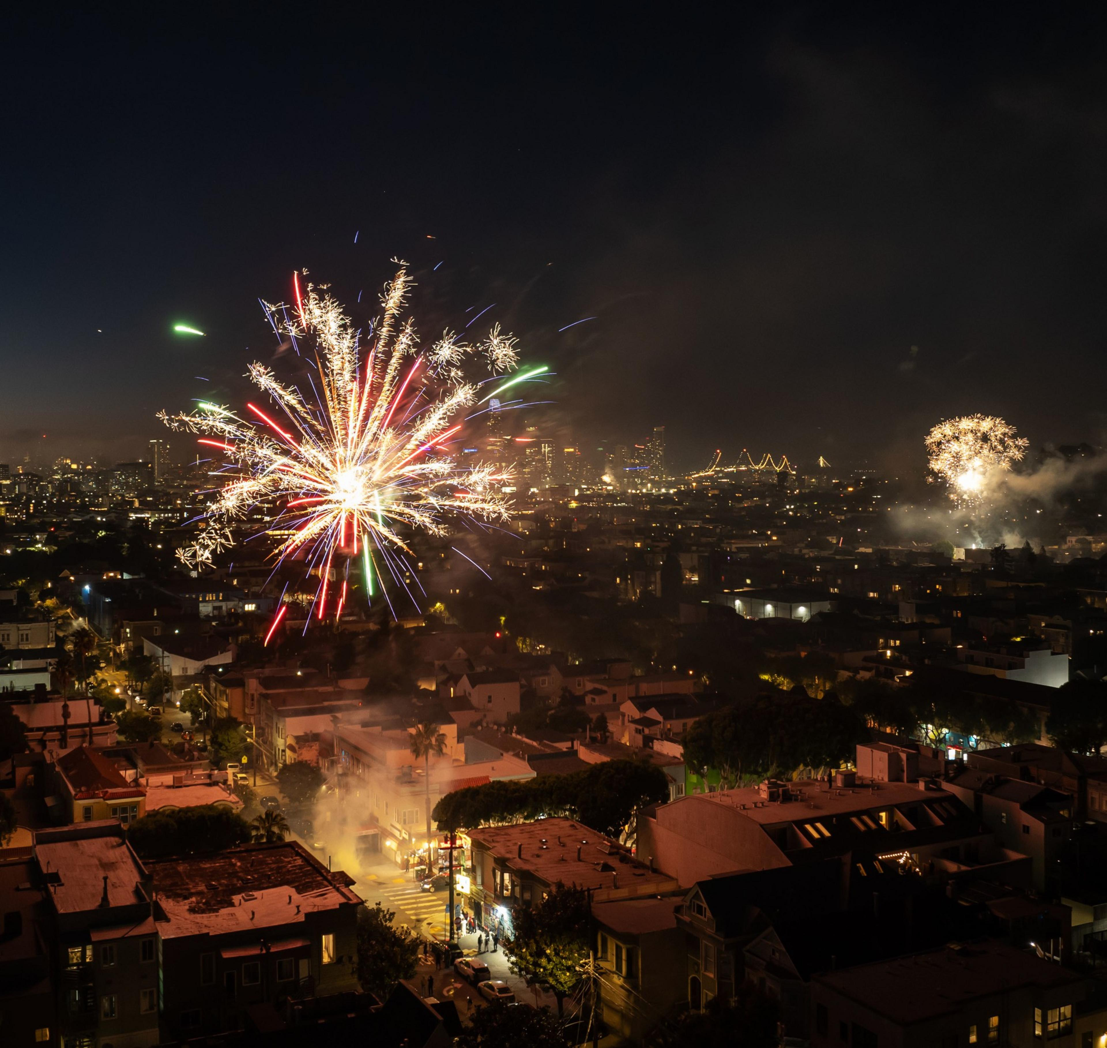 The image shows a nighttime cityscape with vibrant fireworks. Houses and streets are illuminated in the foreground, while a bustling city with bridges is in the background.