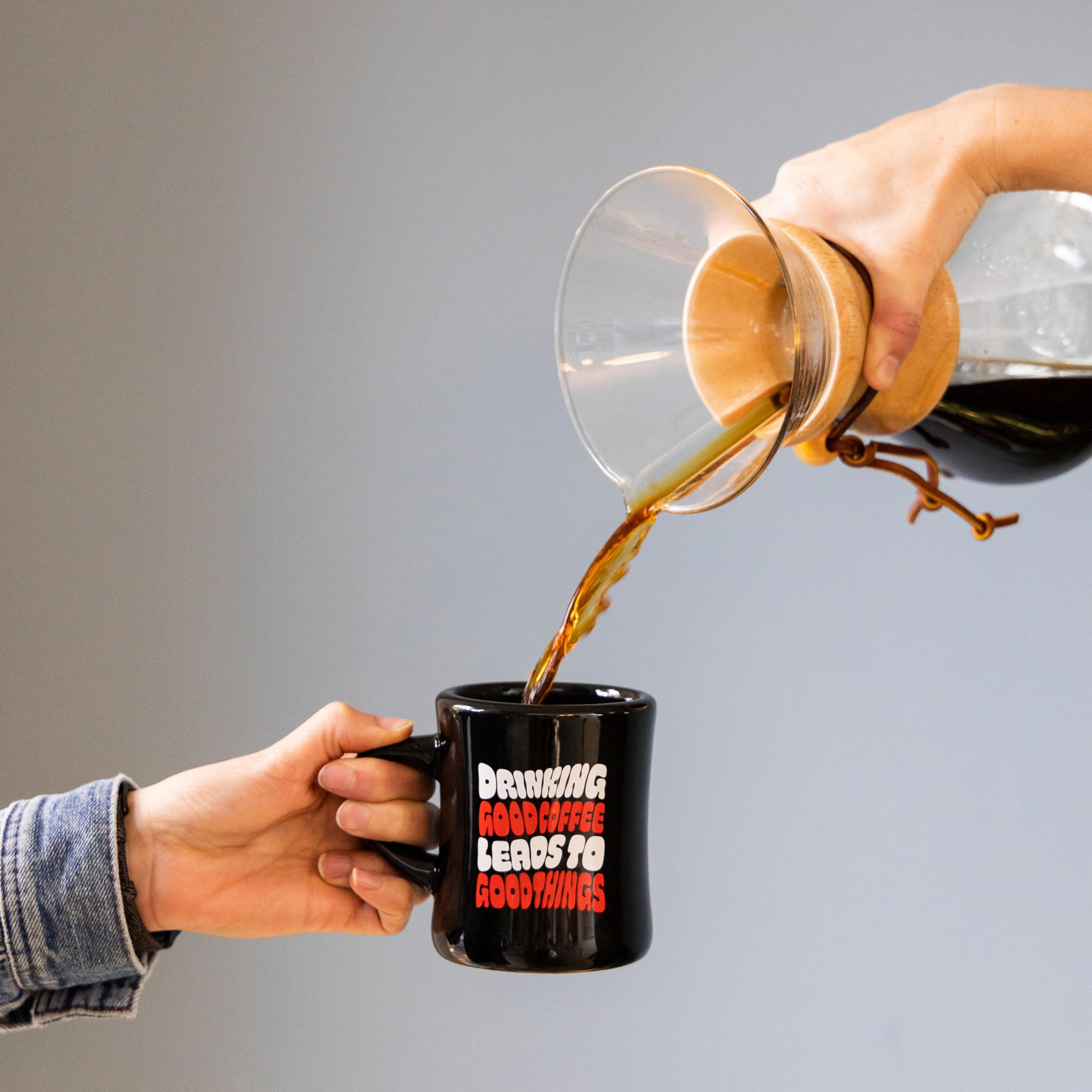 Black coffee pours from a glass Chemex into a black mug held by a hand wearing a denim jacket. The mug reads, "DRINKING GOOD COFFEE LEADS TO GOOD THINGS."