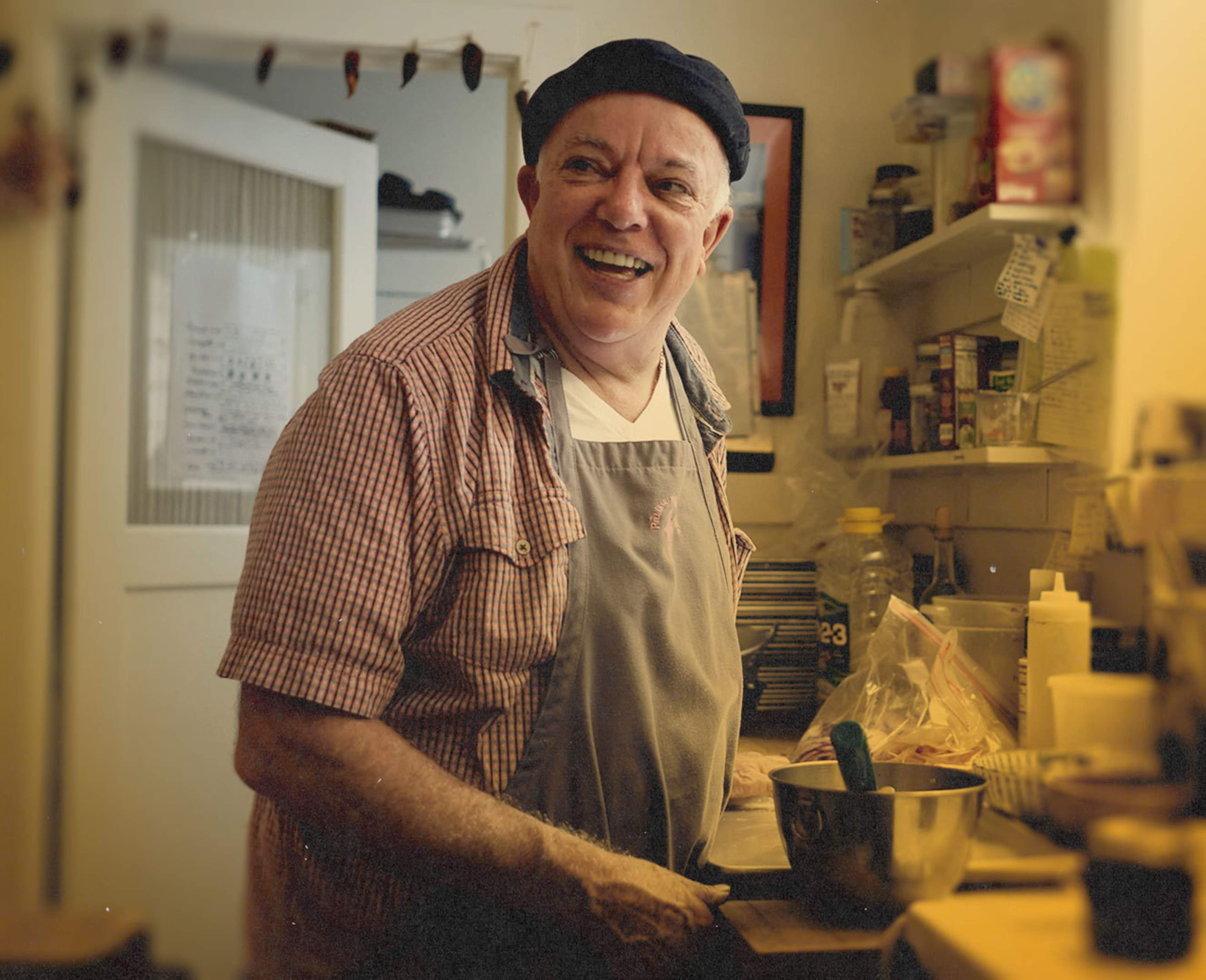A smiling man wearing a black beret, checkered shirt, and apron stands in a cozy kitchen beside a mixing bowl and various cooking items.