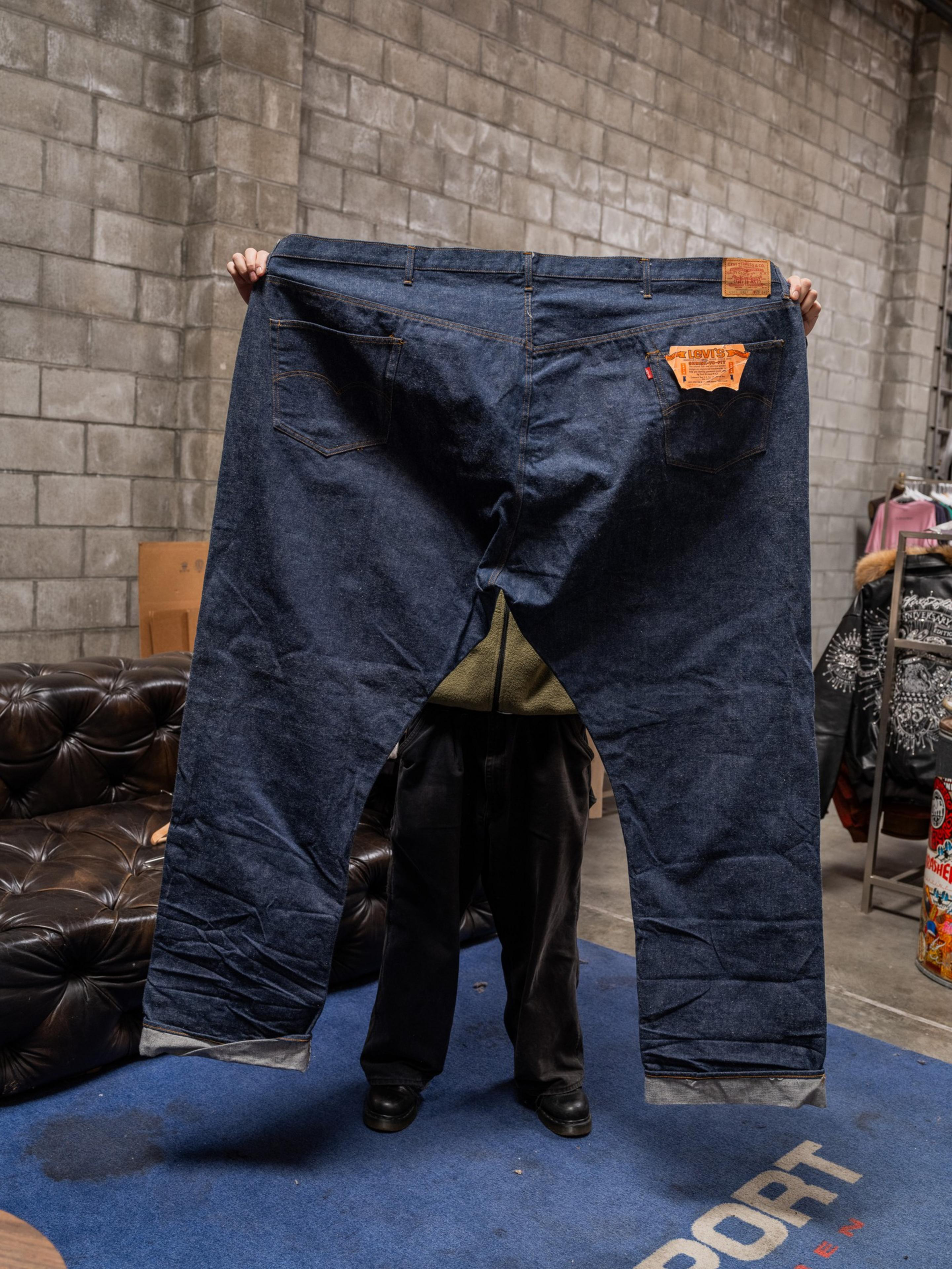 A person is holding up an enormous pair of blue jeans, wide enough to cover their entire body from view. The jeans appear comically oversized.