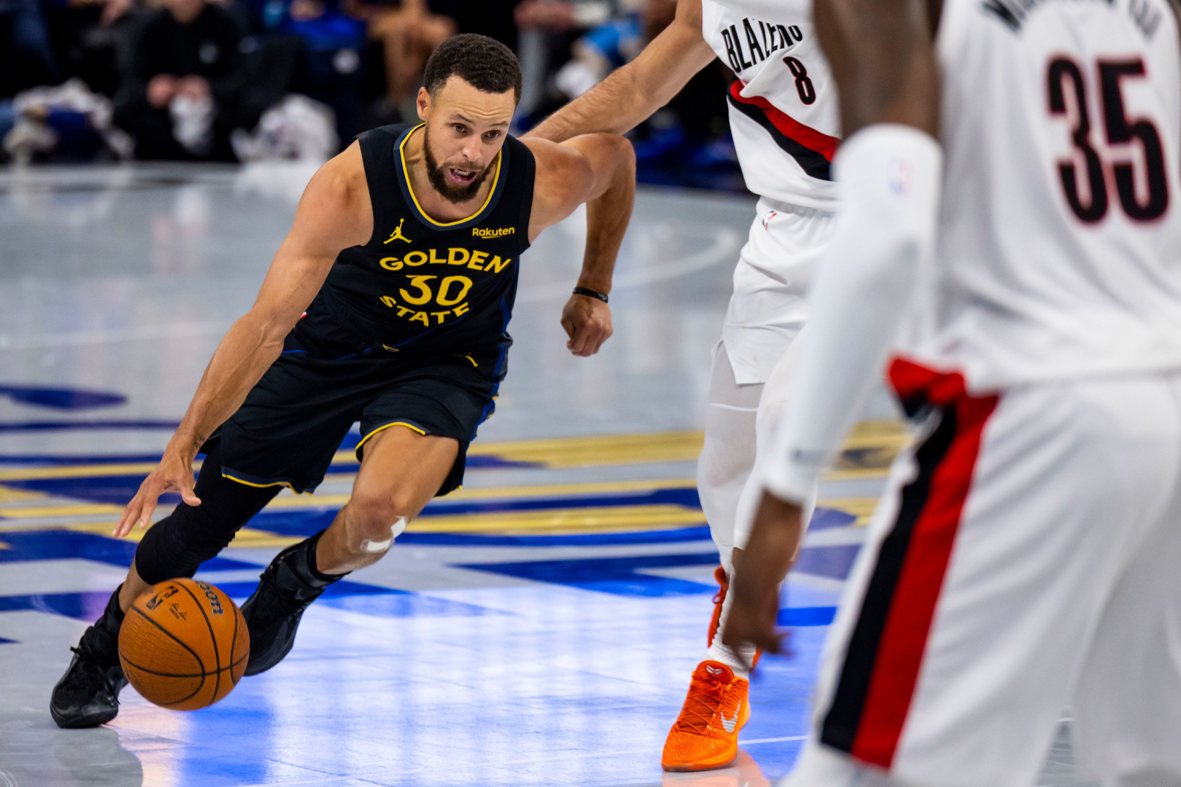 A basketball player in a Golden State Warriors jersey dribbles the ball while two defenders in Portland Trail Blazers uniforms guard him.
