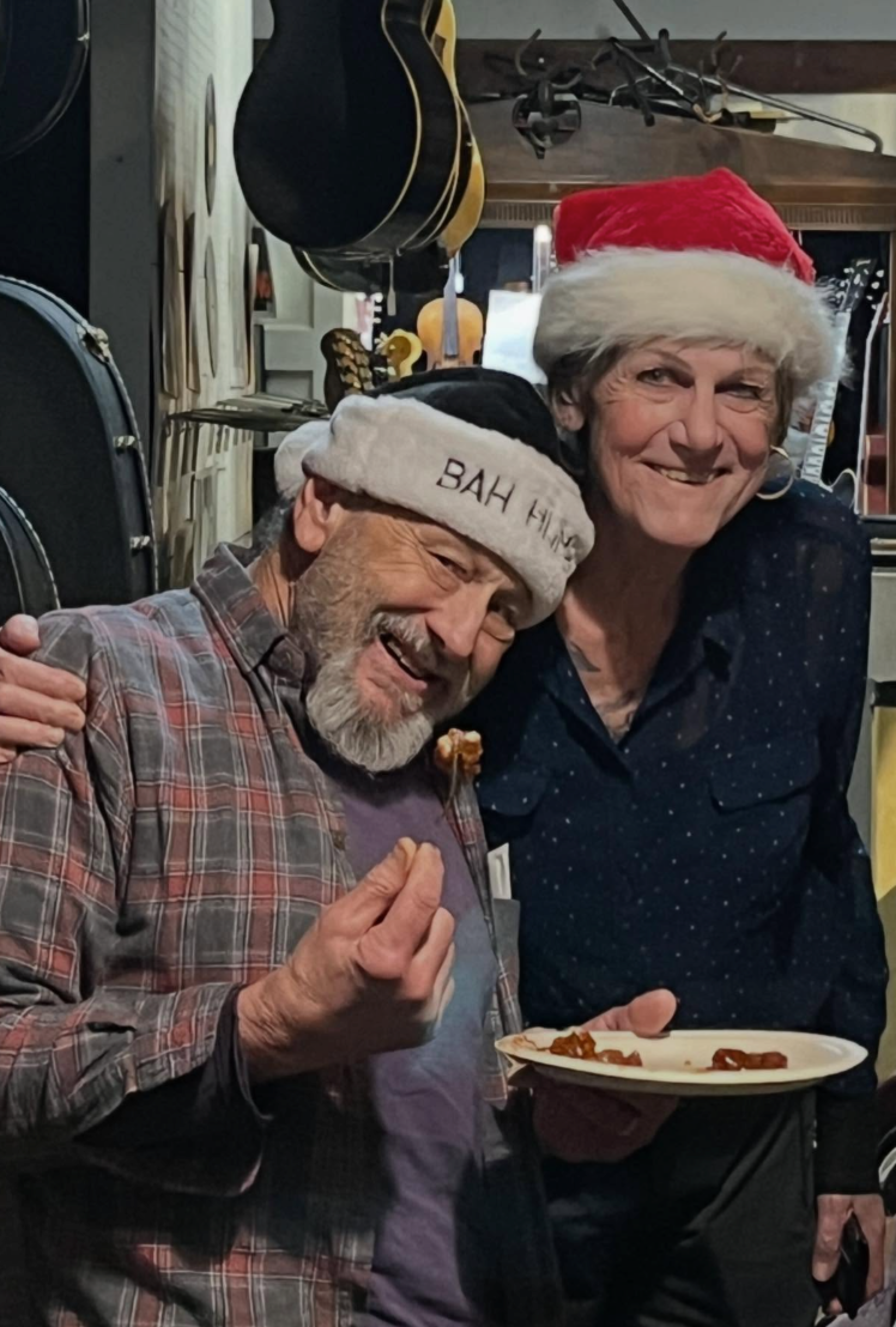 A smiling man and woman wearing Santa hats share a moment; the man holds a bite of food, and the woman holds a plate with food on it.