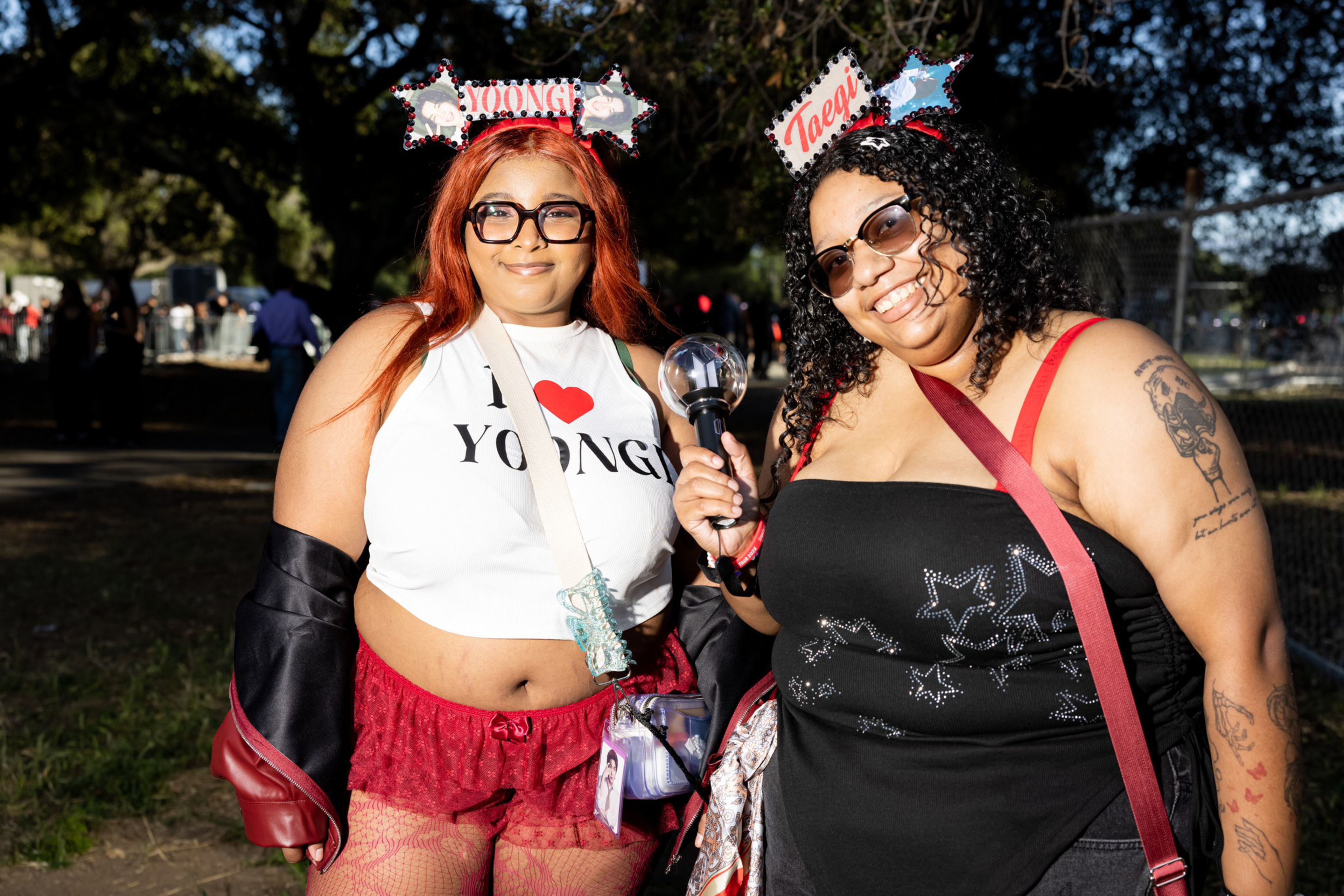 Two women smile outdoors wearing glasses and headbands labeled “Yoongi” and “Taegi,” with one holding a lightstick and the other wearing a shirt that says “I ♥ Yoongi.”