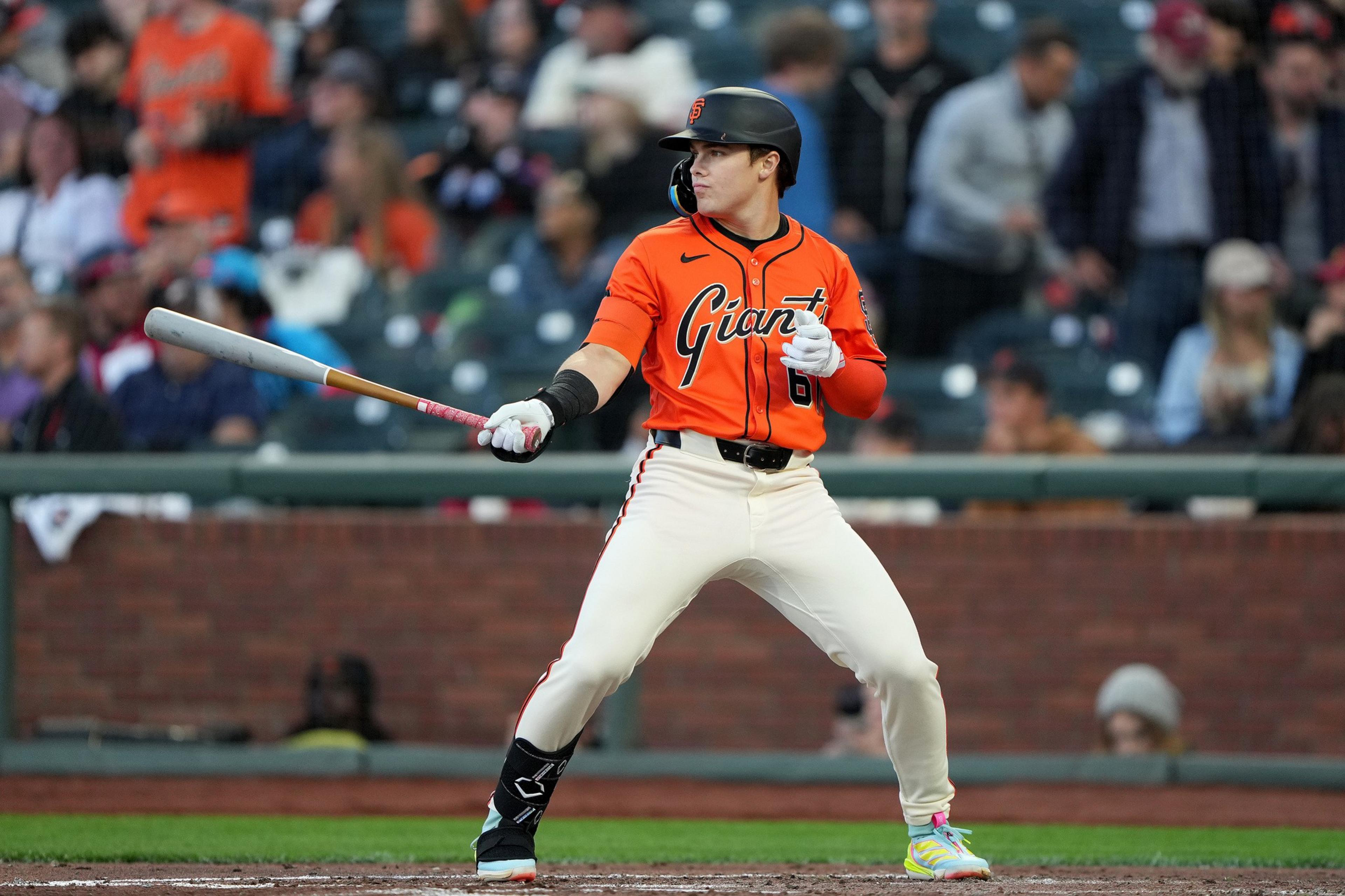 A baseball player in an orange Giants jersey stands ready to bat, holding the bat out in front, with spectators blurred in the background.