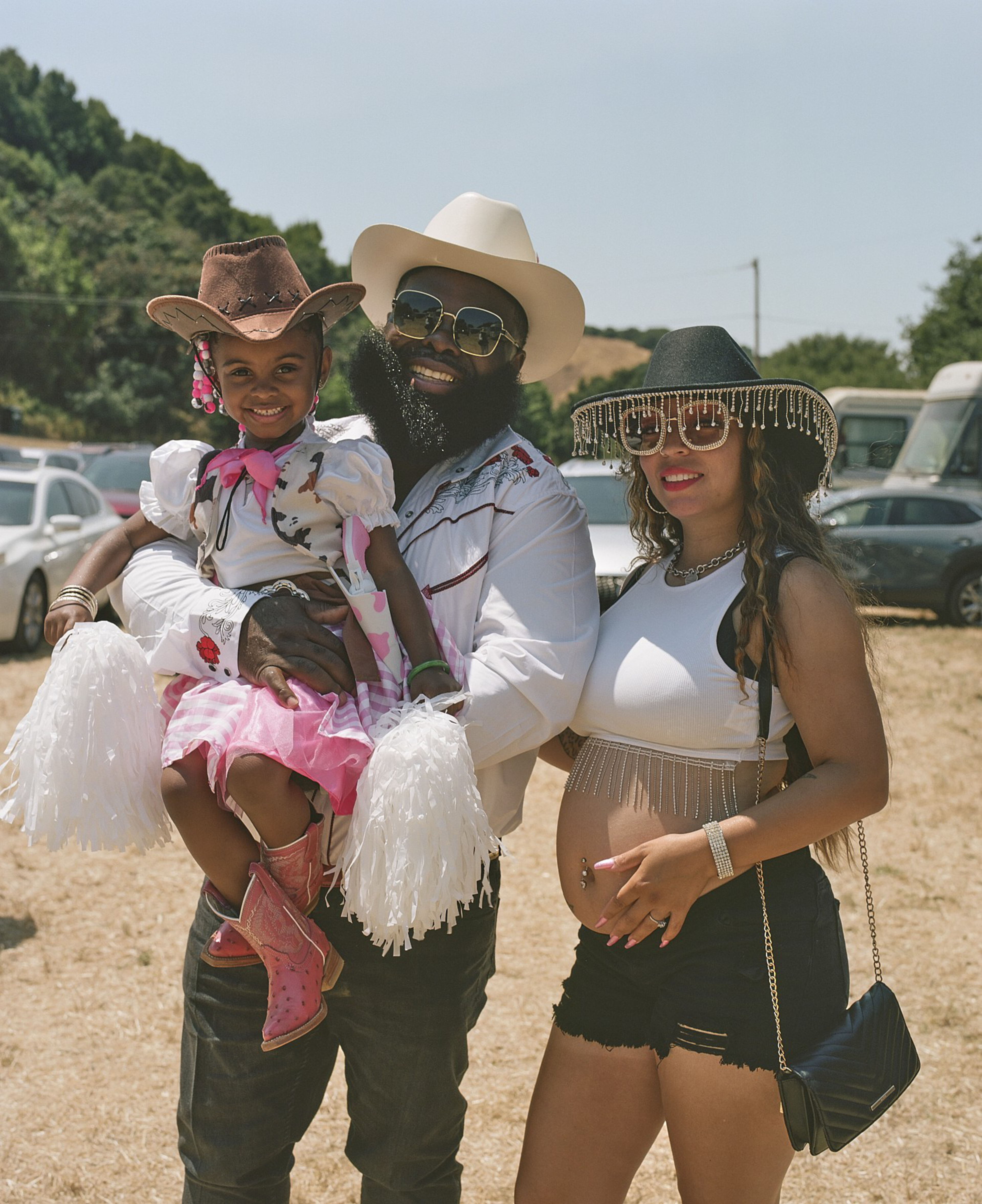 A family poses outdoors. The father holds a smiling child in cowgirl attire. The mother stands beside them, wearing a fringed hat, her hand on her belly.